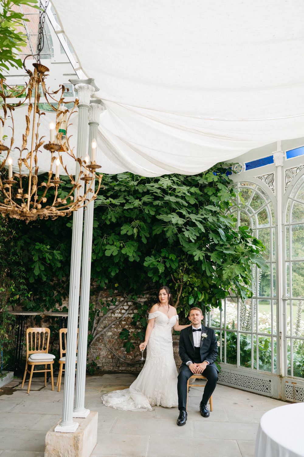 A couple sits together at their elegant wedding venue under a white gazebo with greenery and chandeliers.
