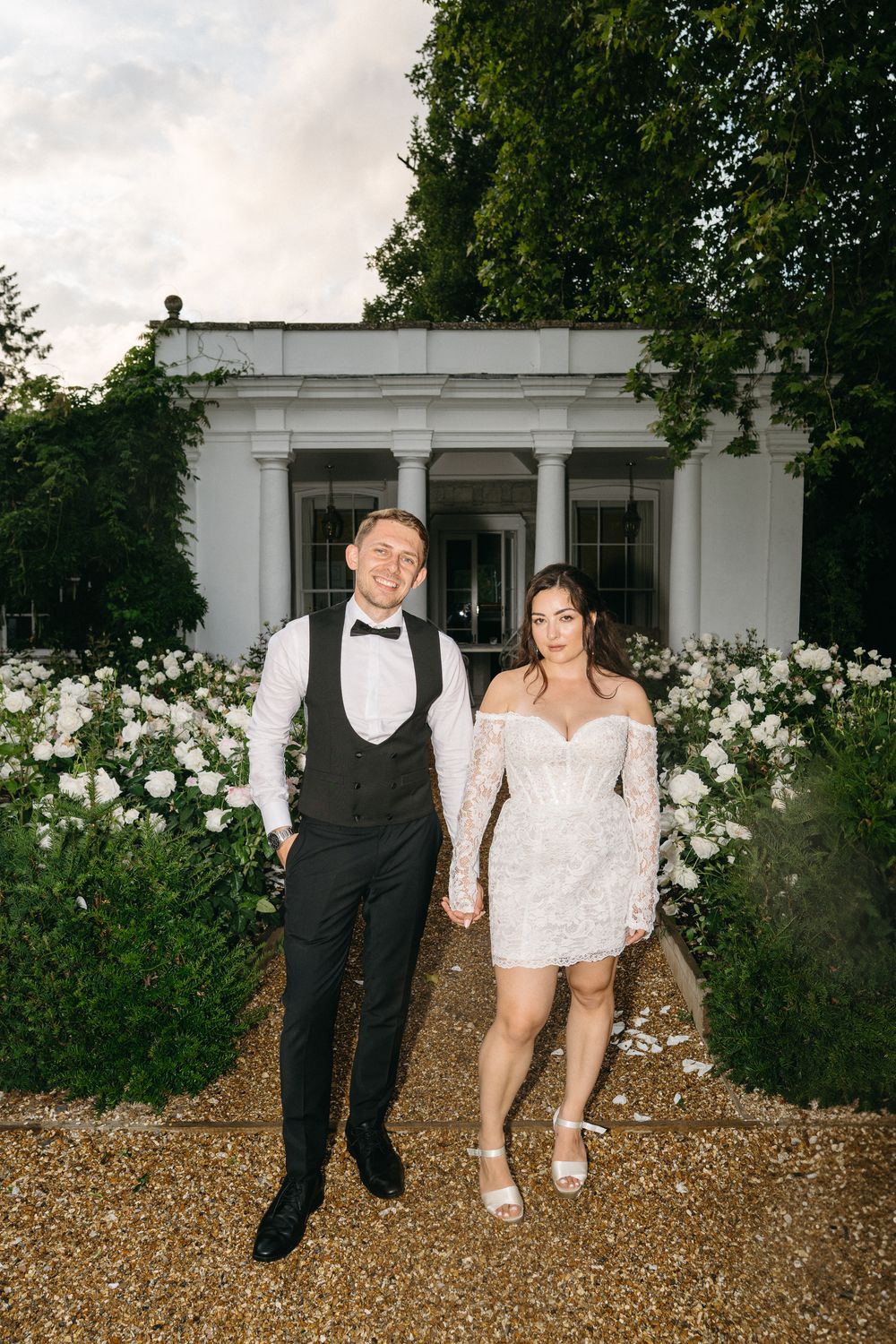 A playful wedding couple sitting on gravel ground with white roses and a building facade in background.