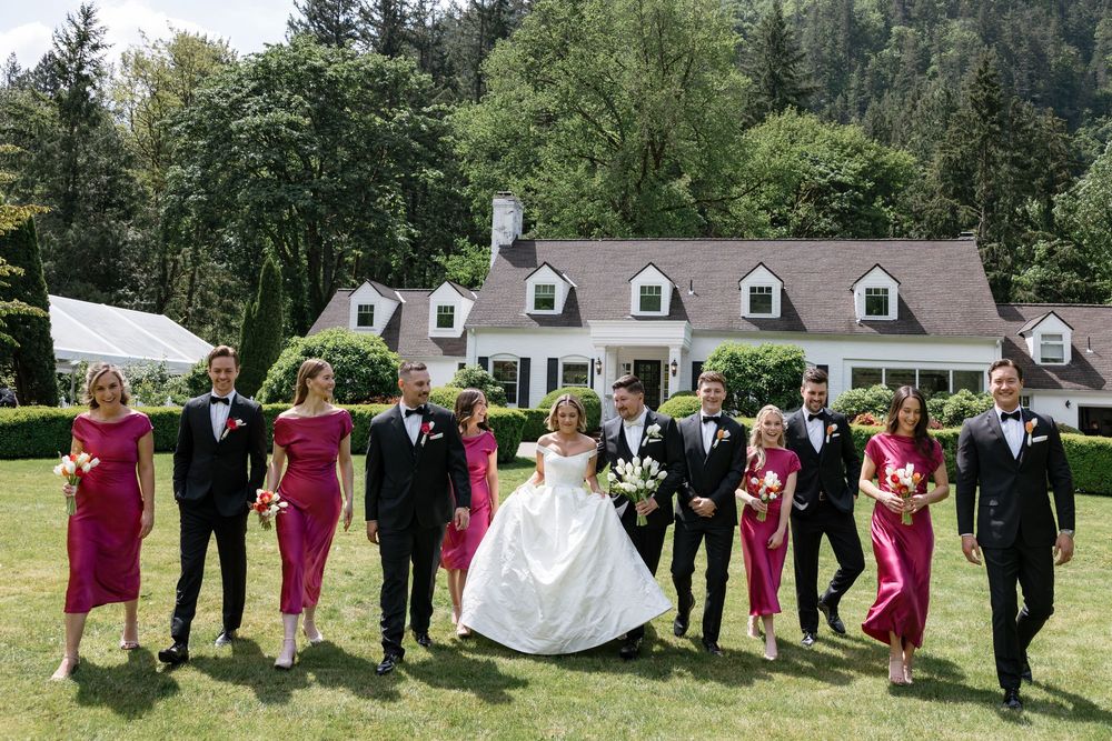 Wedding party poses outdoors in burgundy and black formal attire on a green lawn near white buildings.