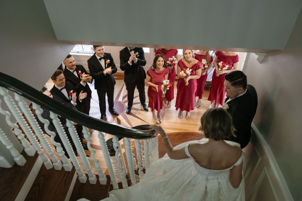 Wedding party gathers on staircase with bridesmaids in red dresses and groomsmen in black suits for photos.