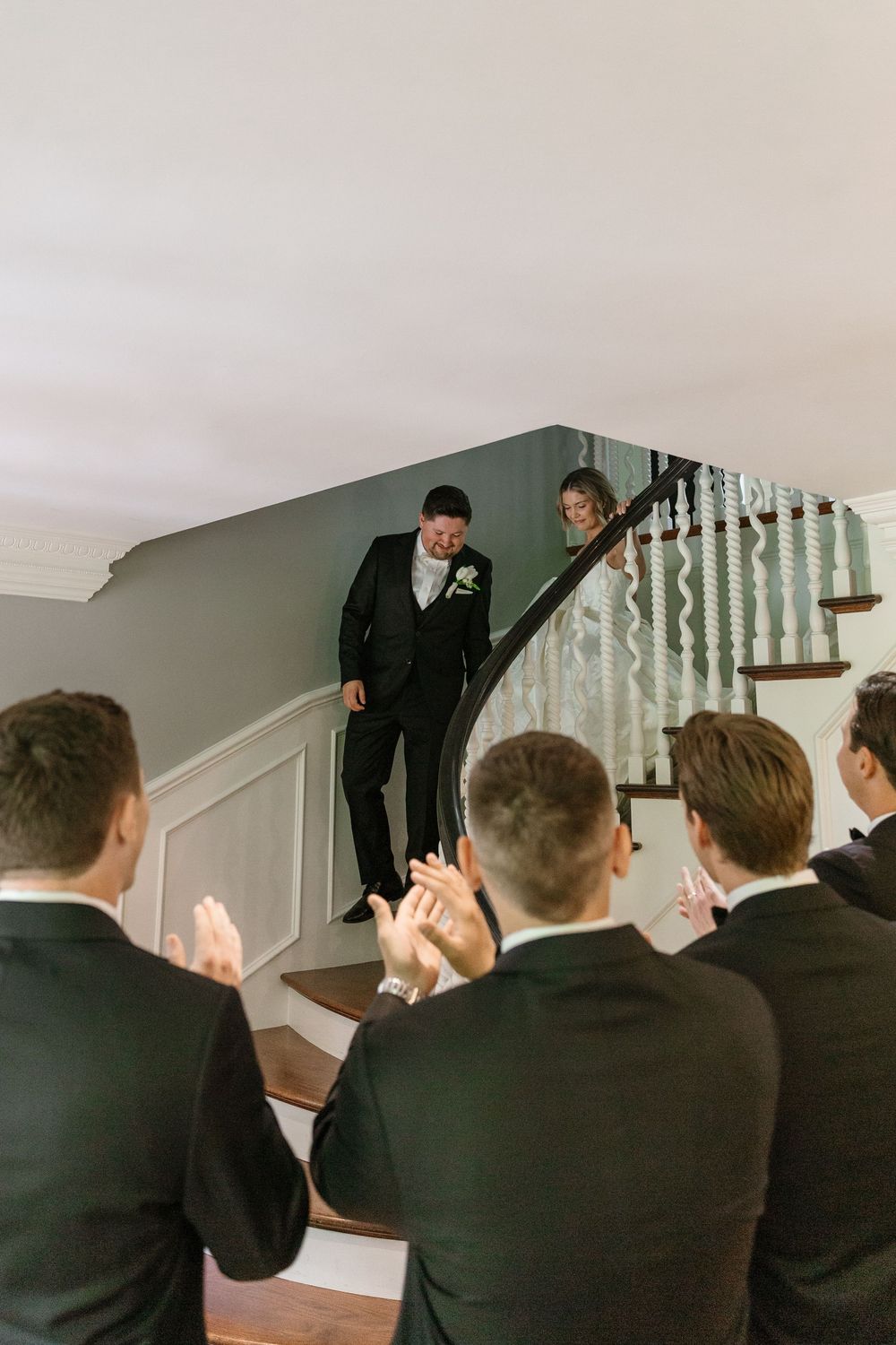 Wedding party members in black suits applaud as couple descends elegant white staircase during celebration moment.