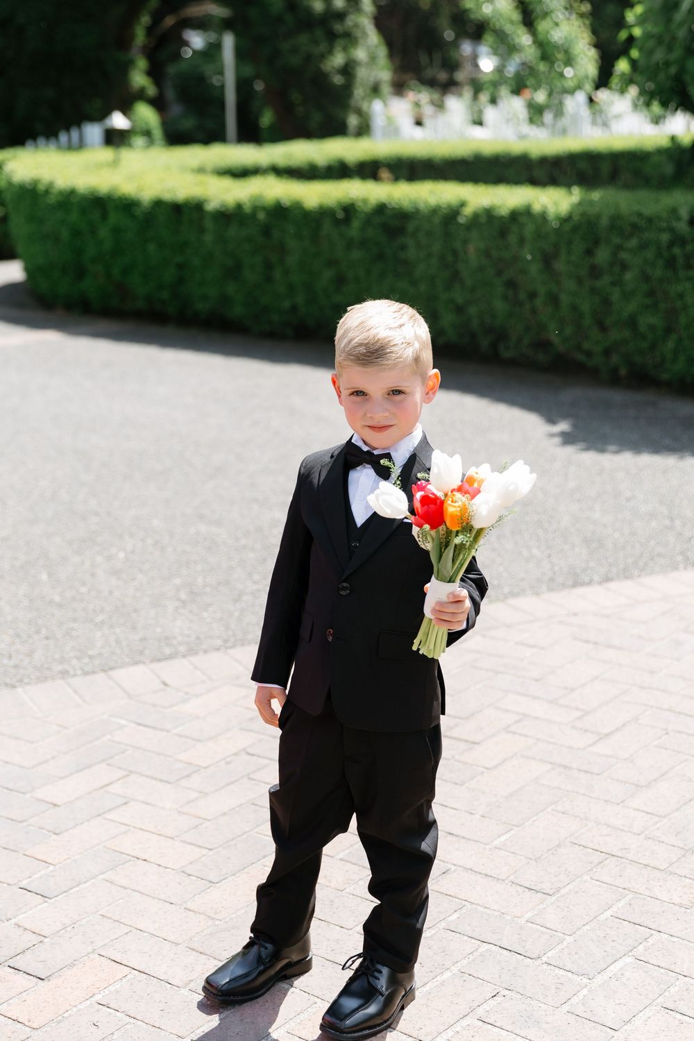 A young ring bearer in a black tuxedo with orange and white floral boutonniere stands on stone pavement.