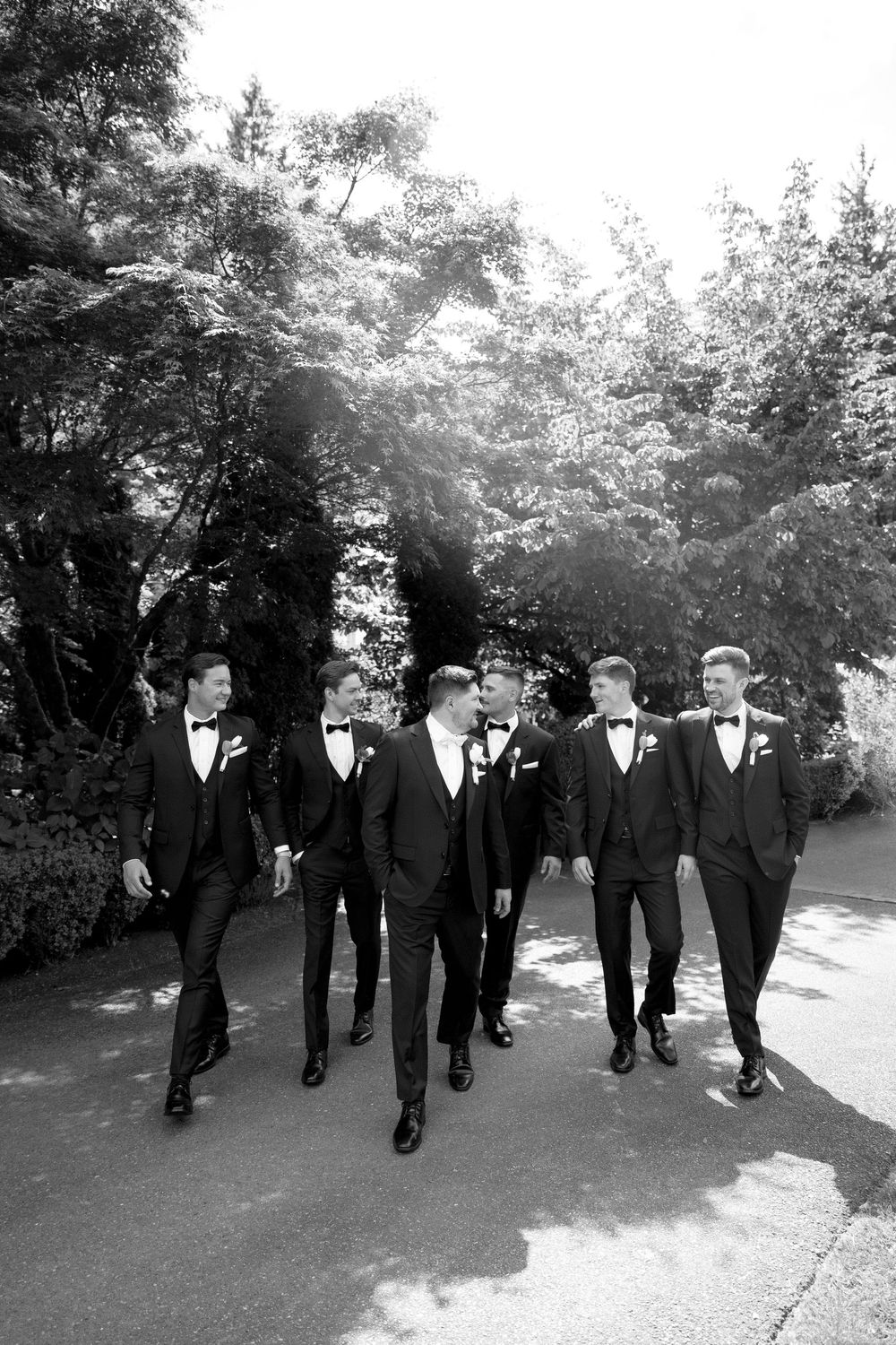 A group of groomsmen in black suits walk together on a tree-lined path in black and white.