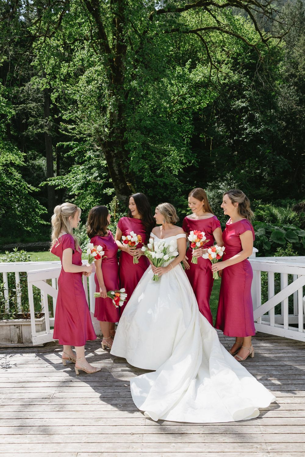Bridesmaids in burgundy dresses pose with bride in white gown on a wooden deck surrounded by lush greenery.