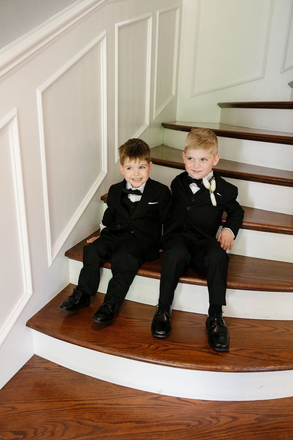 Two young boys in formal black suits sit together on wooden stairs with white paneled walls.