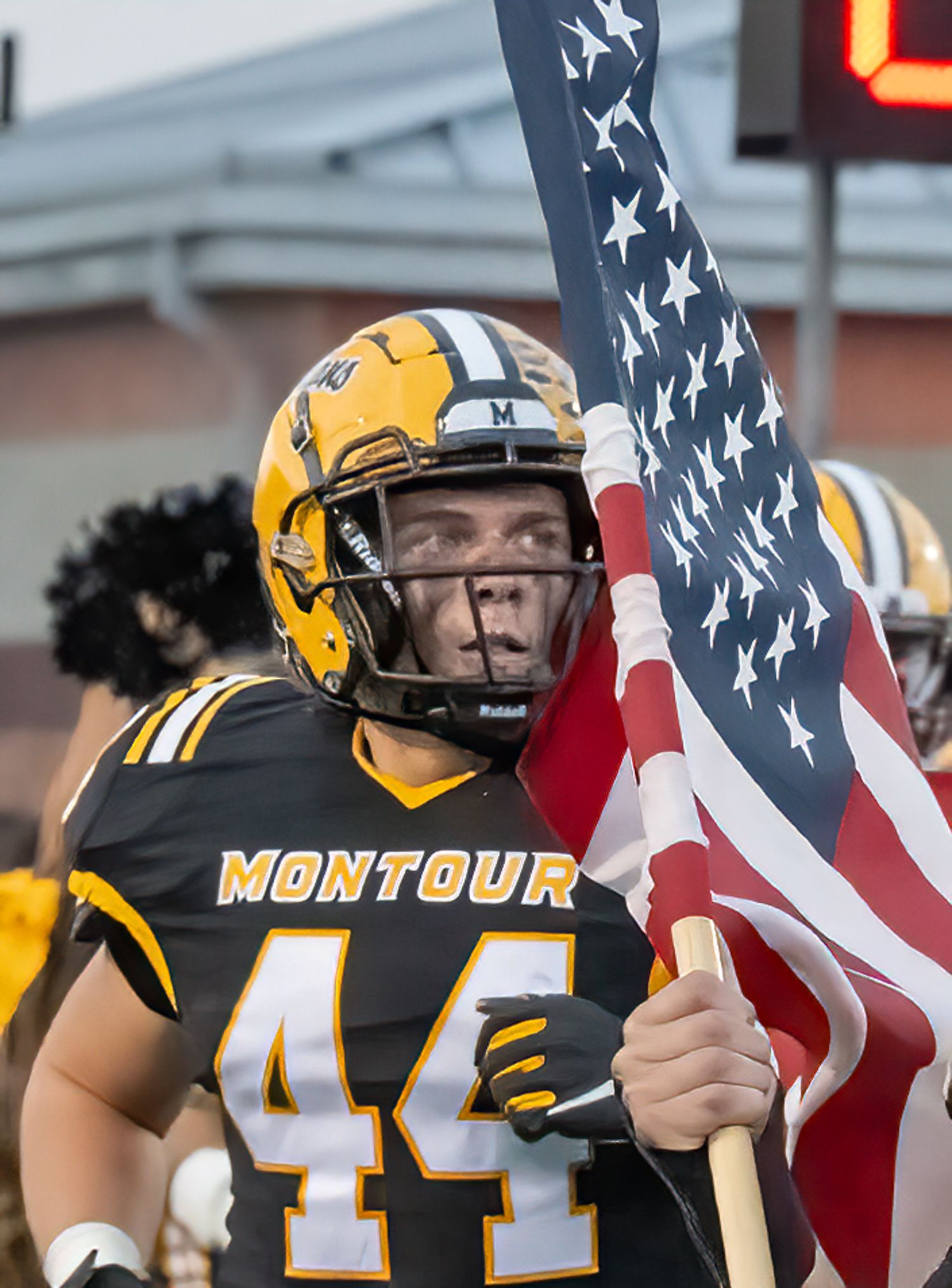 High school football players in black and yellow uniforms stand together with an American flag during a game.