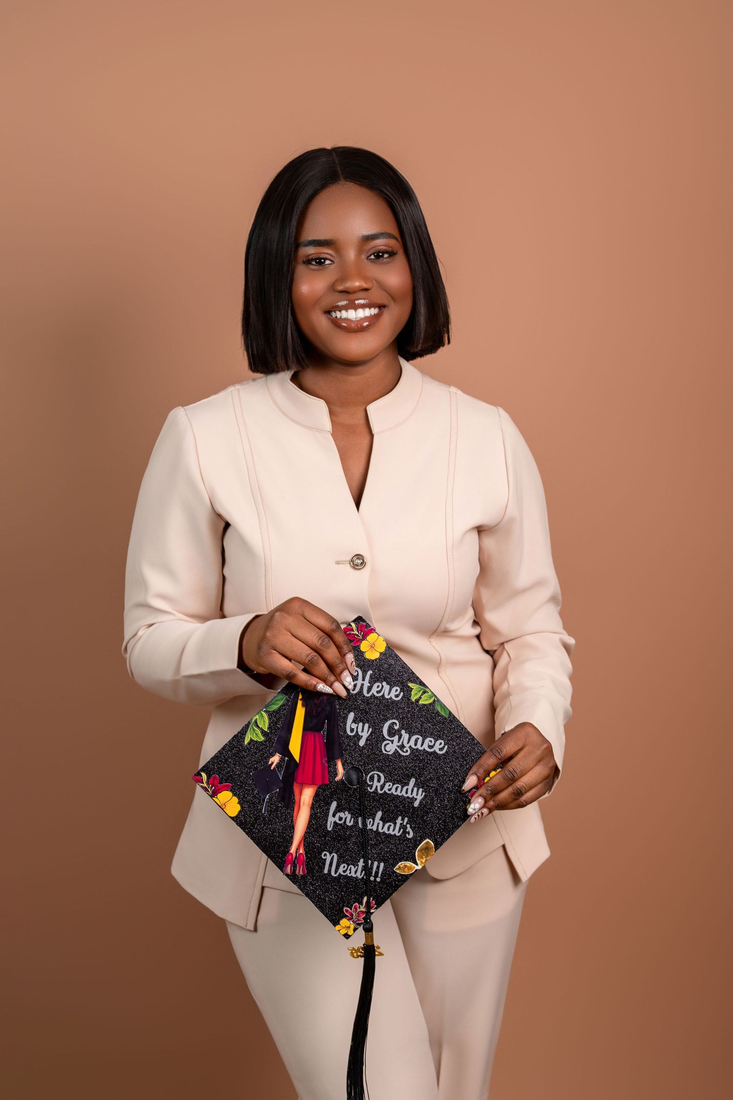Graduate in cream suit holds decorated graduation cap against brown backdrop.