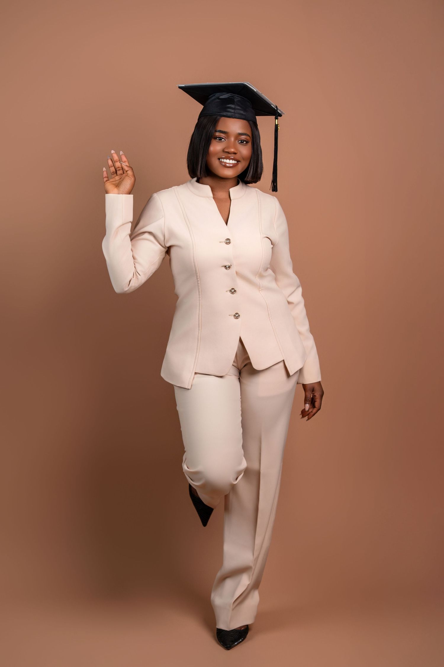 Graduate in cream suit raises hand while posing in graduation cap.