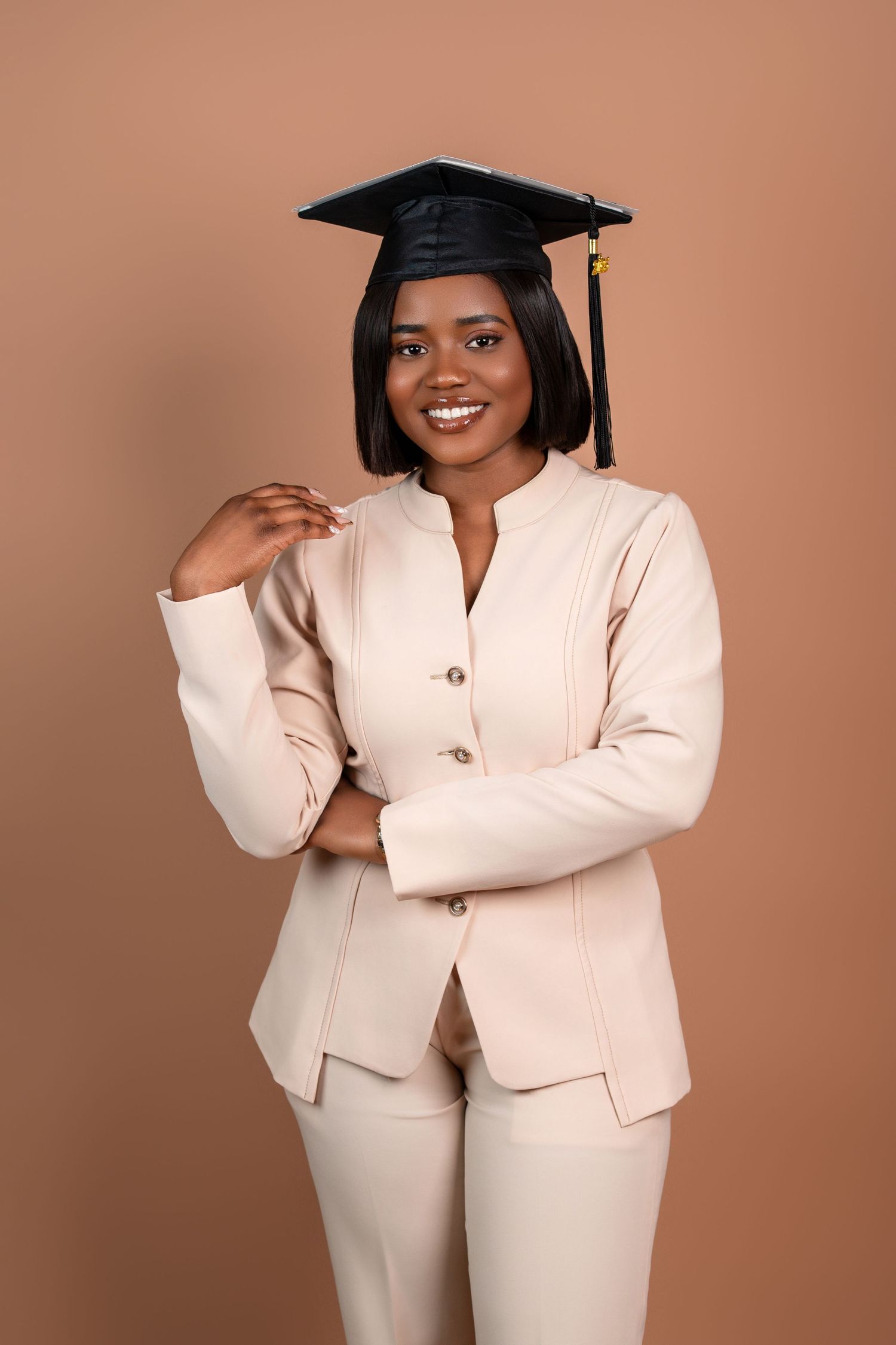 Graduate in cream pantsuit and graduation cap poses against brown background.