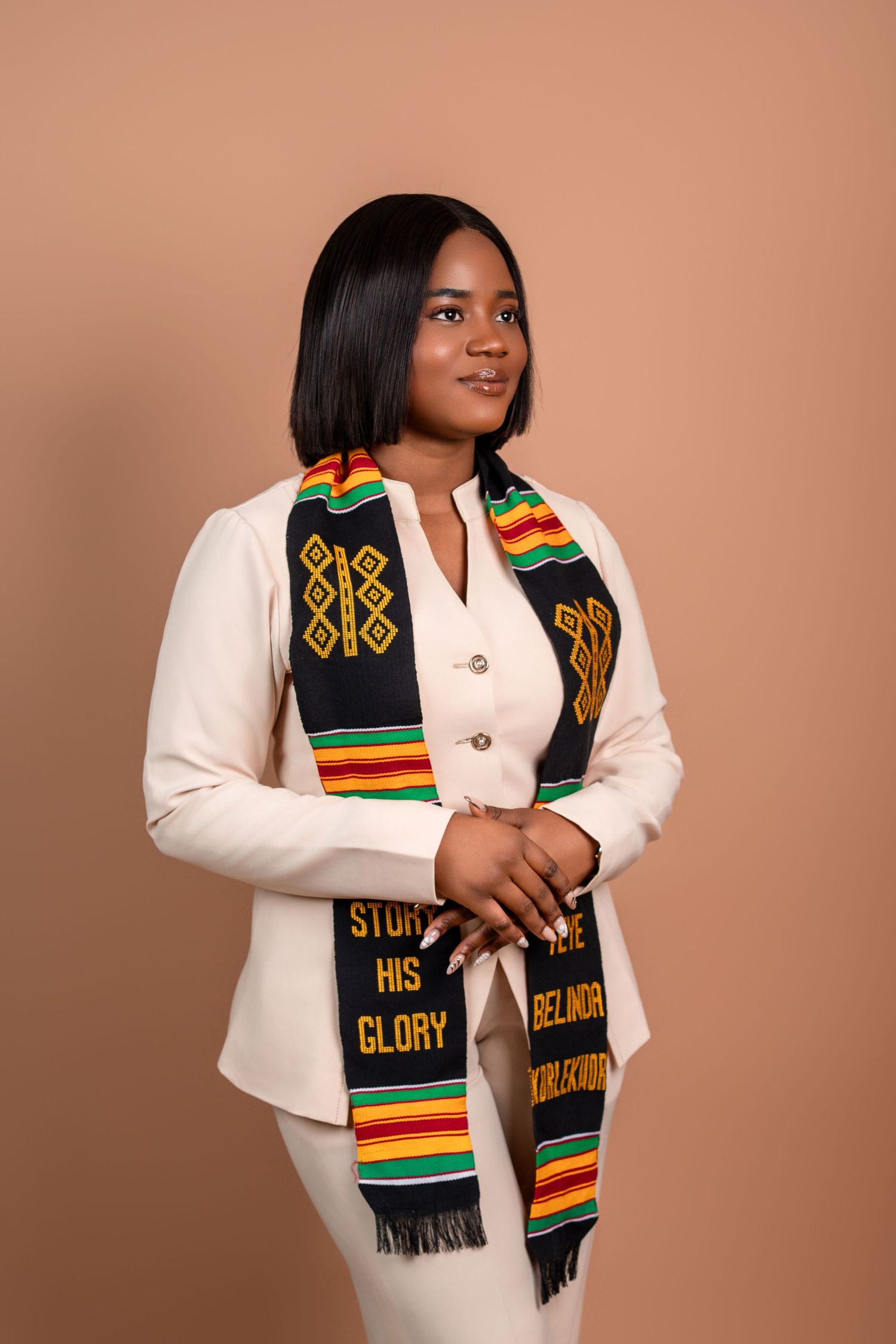 Graduate wearing cream suit and traditional kente stole poses for portrait.