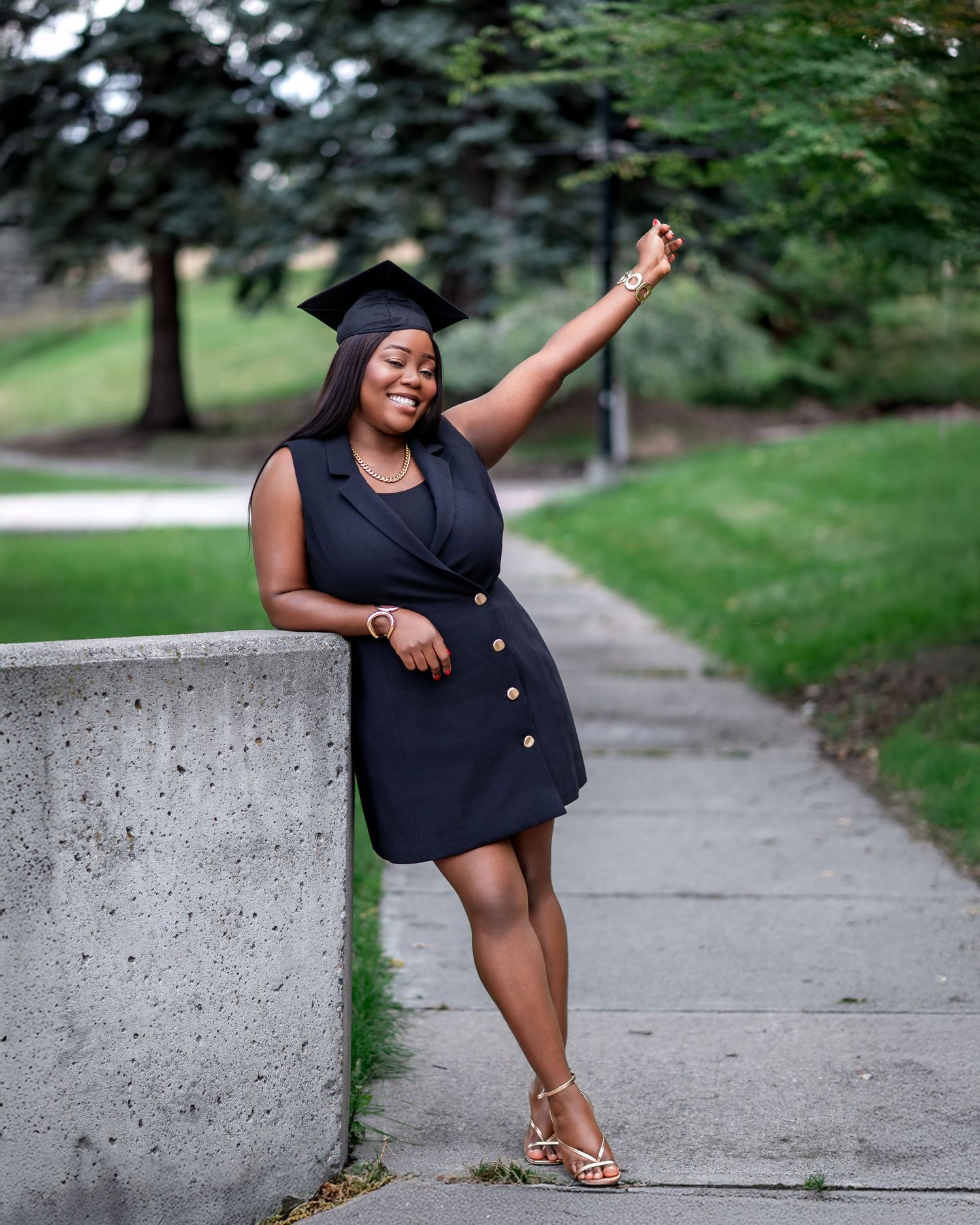 Graduate in black dress poses playfully on concrete wall in park setting.