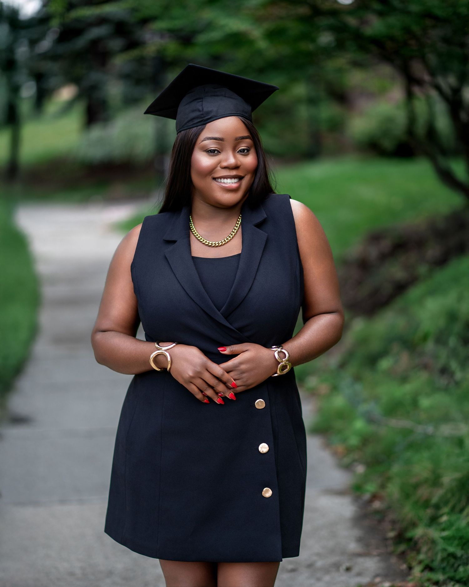 Graduate in black dress and cap poses with jewelry on outdoor path.