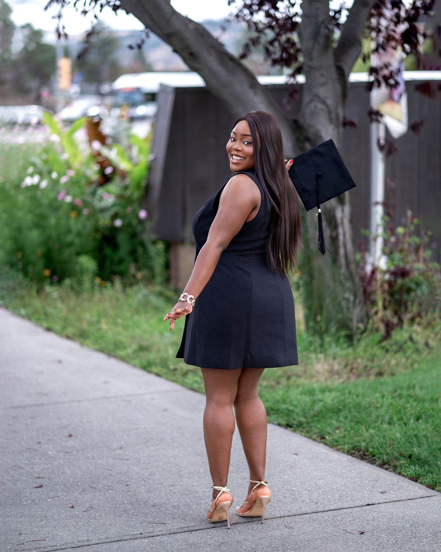 Graduate in black dress strikes elegant poses along campus pathway.