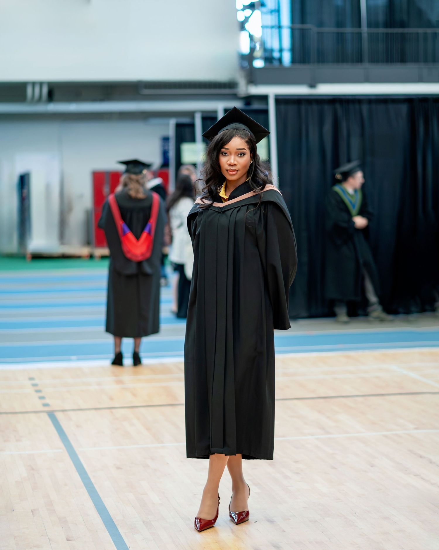 Graduate poses in black academic regalia on indoor gymnasium floor during commencement ceremony.