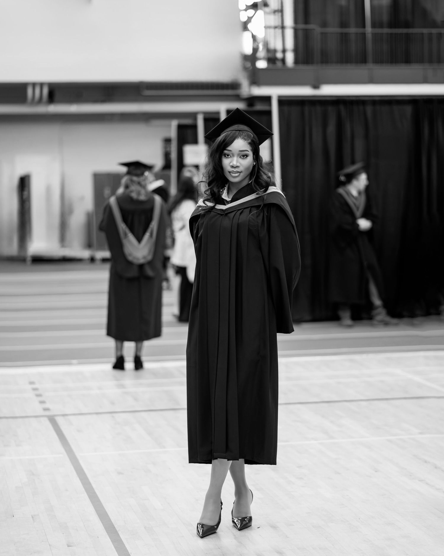 Black and white portrait of graduate in academic regalia posing in gymnasium.