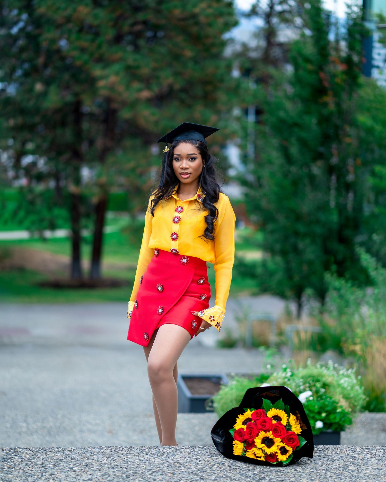 Graduate in yellow blouse and red skirt poses with sunflower bouquet on outdoor path.