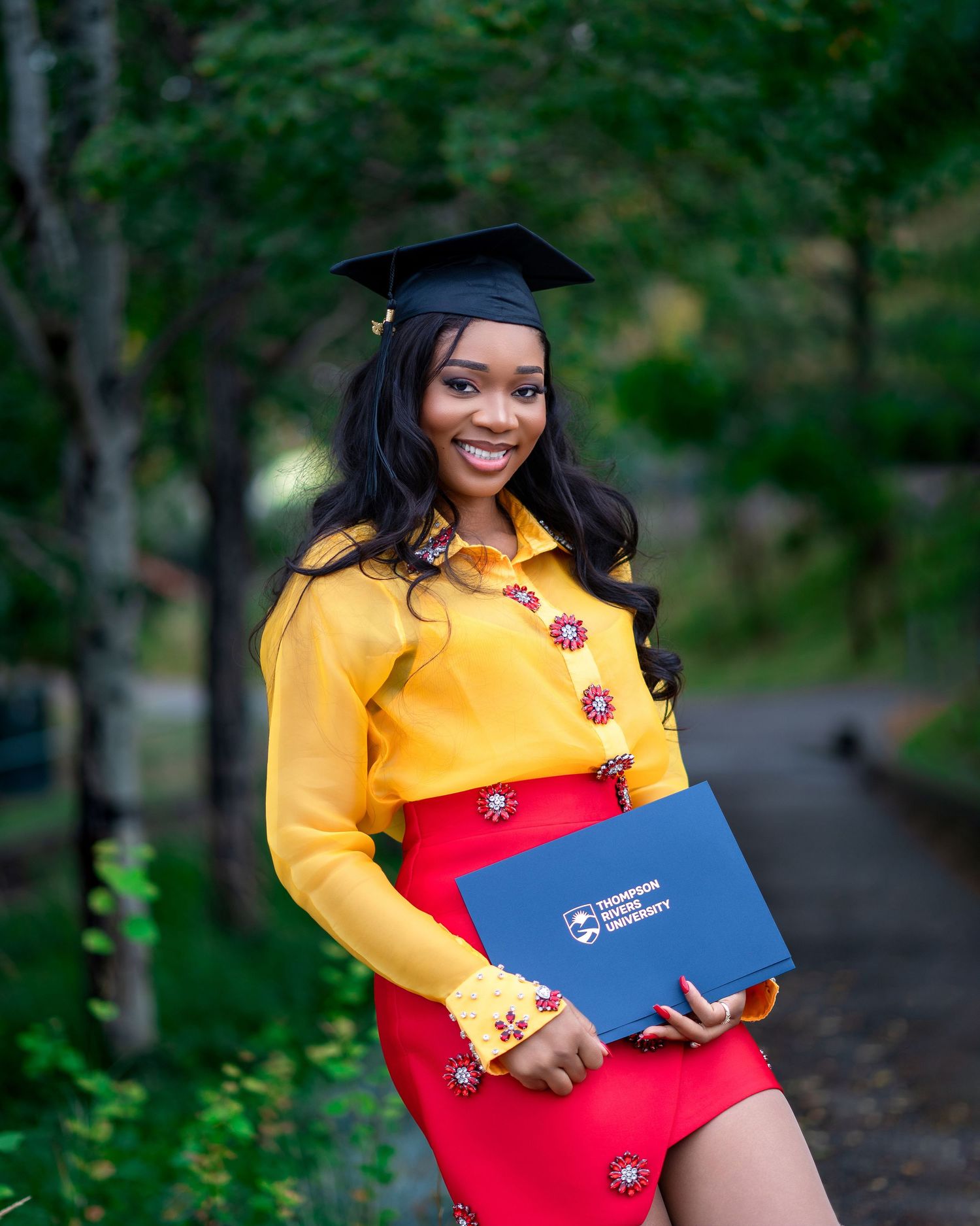 Graduate in yellow top and red skirt holds blue diploma cover during outdoor photo session.