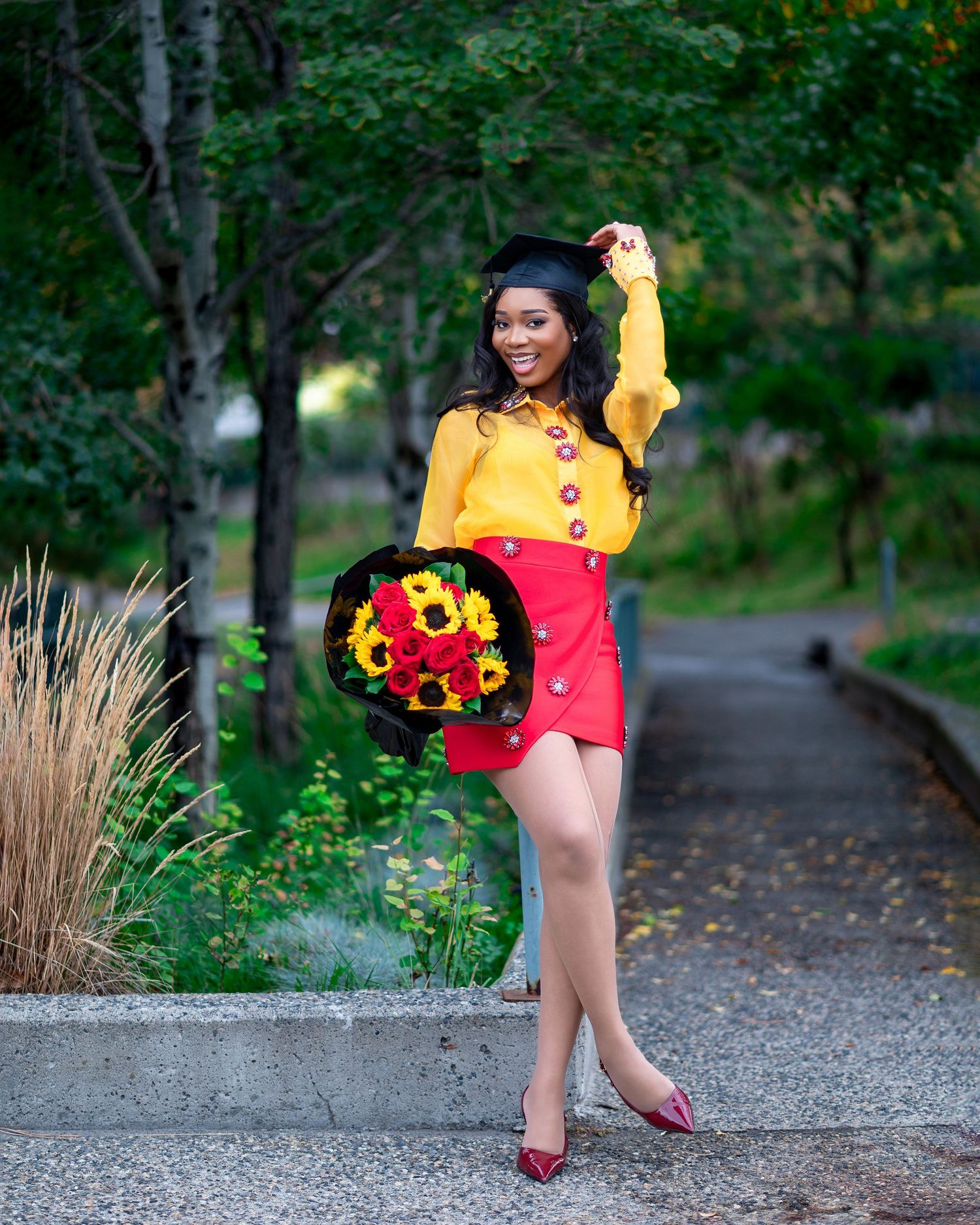 Graduate in yellow blouse and red skirt poses playfully with sunflower bouquet on campus path.