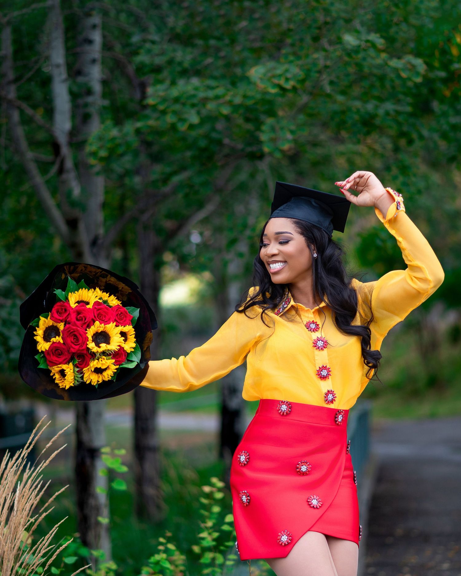 Graduate in vibrant yellow and red outfit celebrates with sunflower bouquet outdoors.