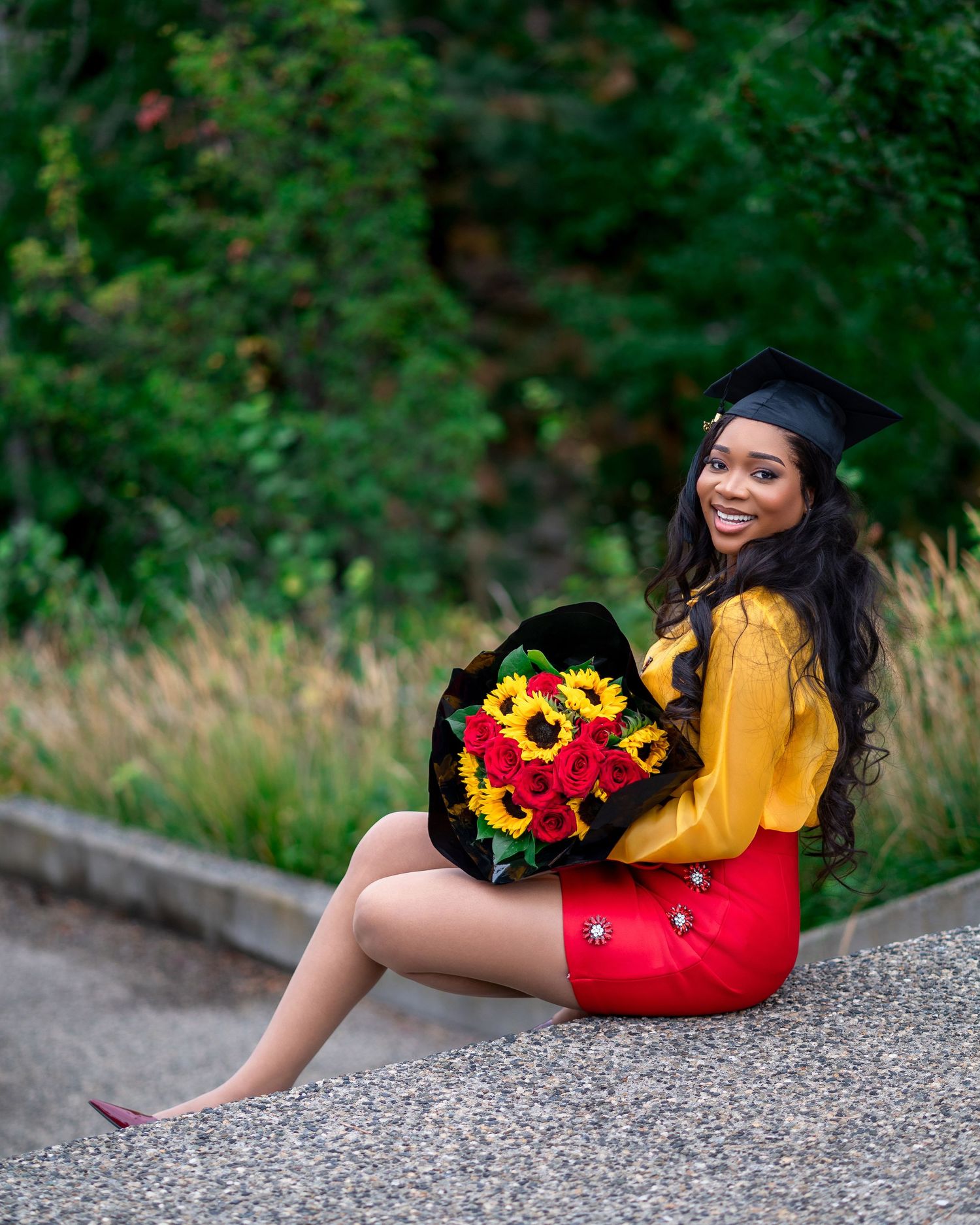 Graduate in yellow and red outfit poses with sunflowers against green foliage background.