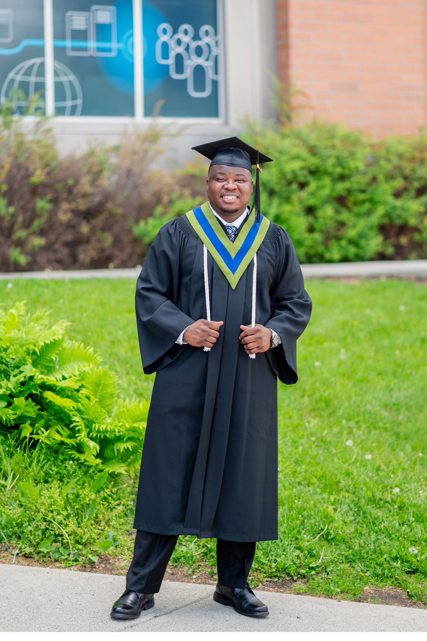 Graduate in black cap and gown with blue and yellow stole stands proudly on campus grounds.