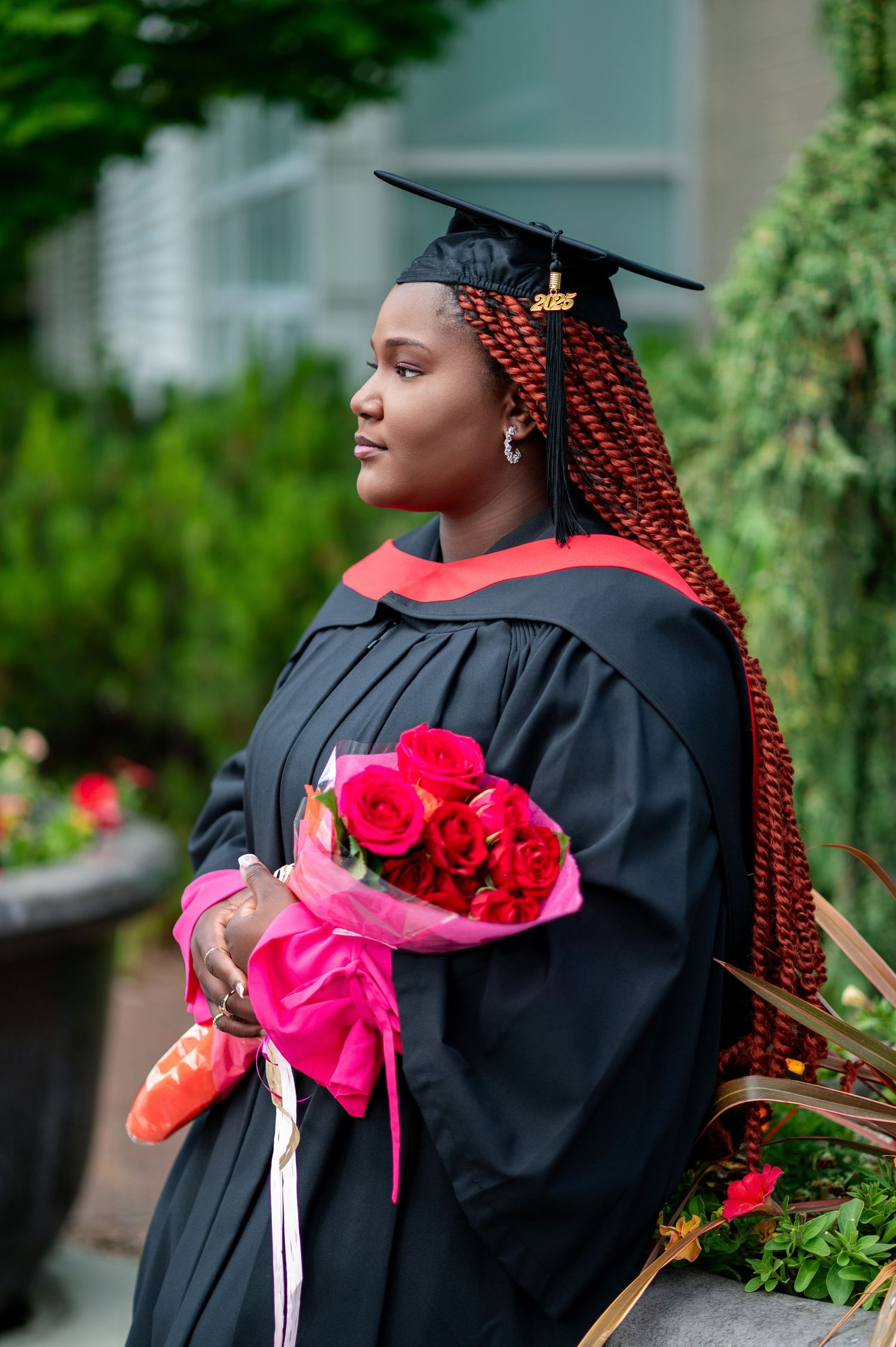 Graduate with braided hair poses in profile while holding red roses outdoors.