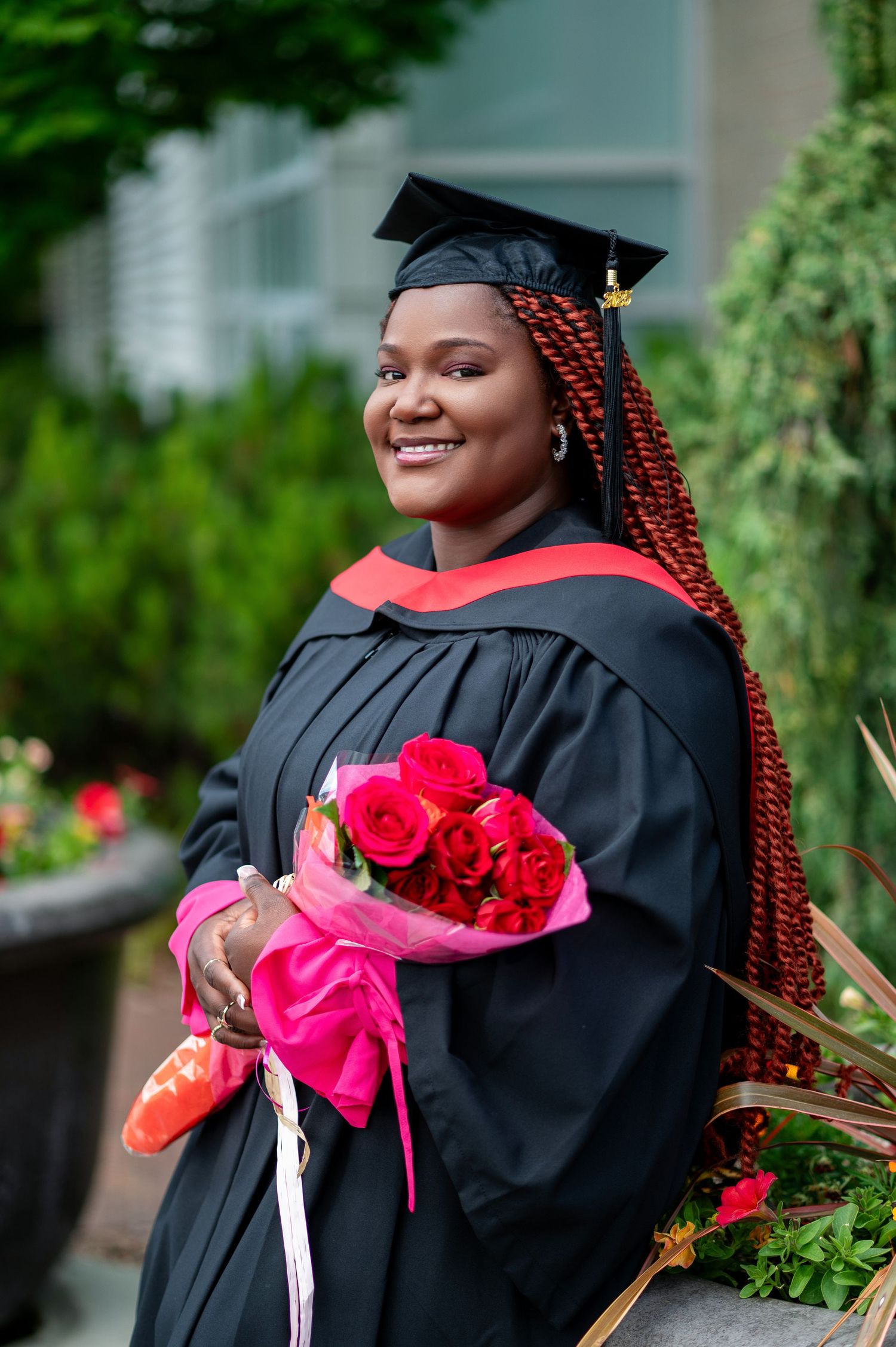 Graduate in black cap and gown holds red roses during outdoor graduation photoshoot.