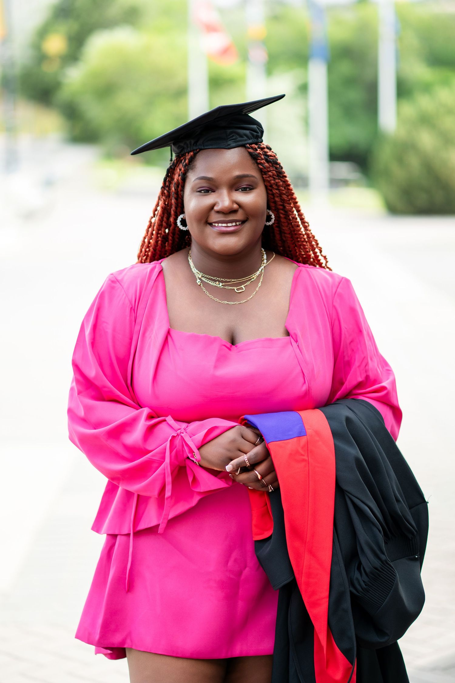 Graduate wearing bright pink dress and academic regalia smiles outdoors.