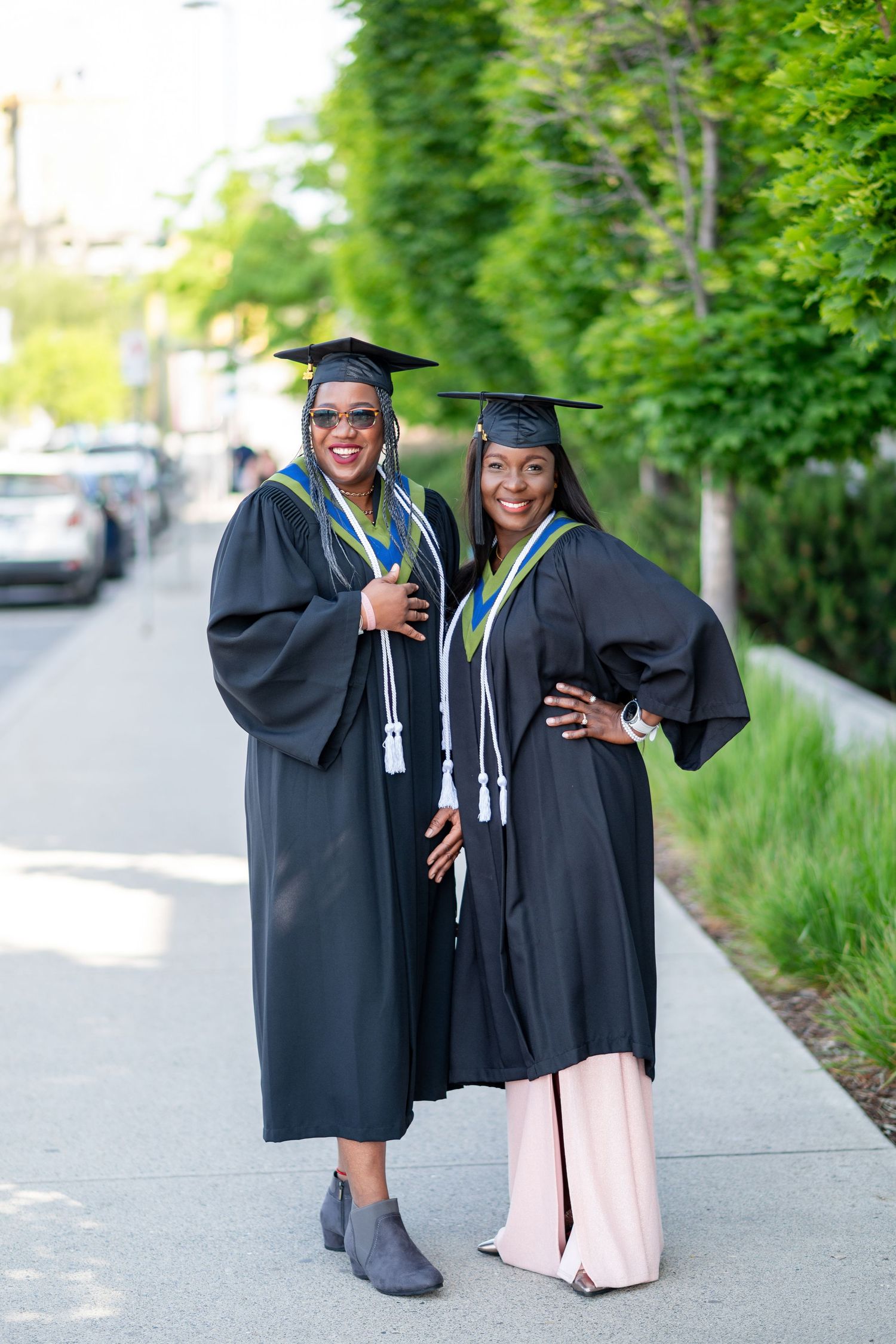 Two graduates in black caps and gowns celebrate together outdoors with joyful expressions.