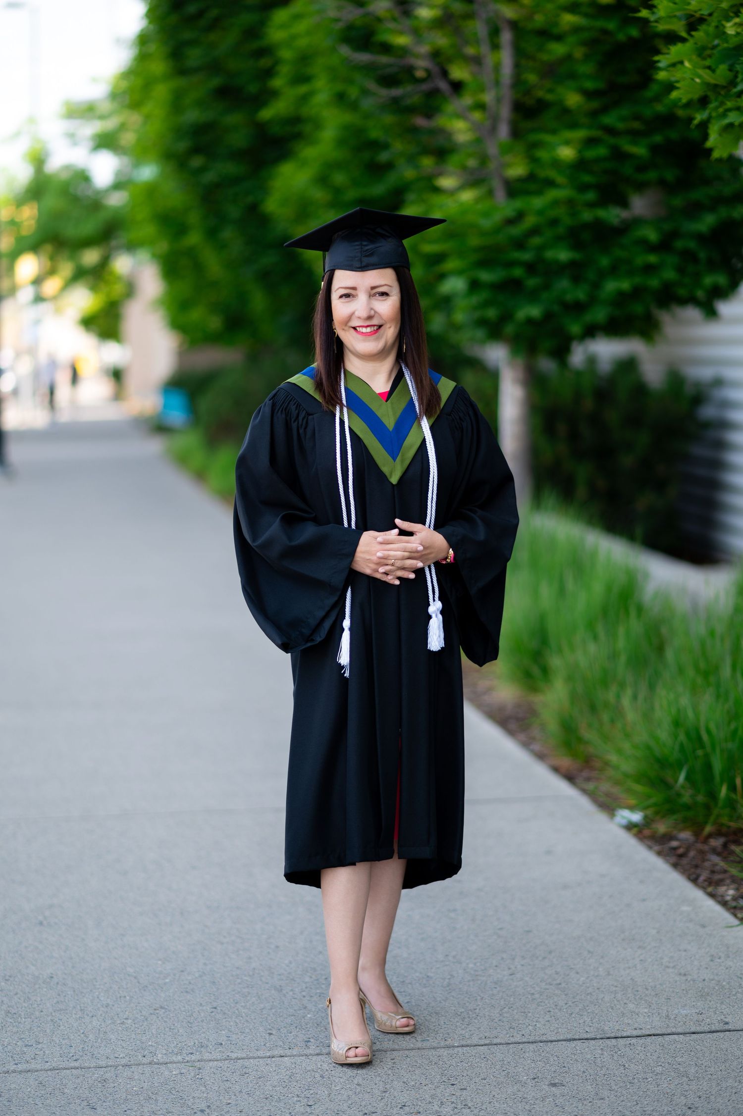 Graduate in academic regalia poses gracefully on outdoor pathway with green foliage backdrop.