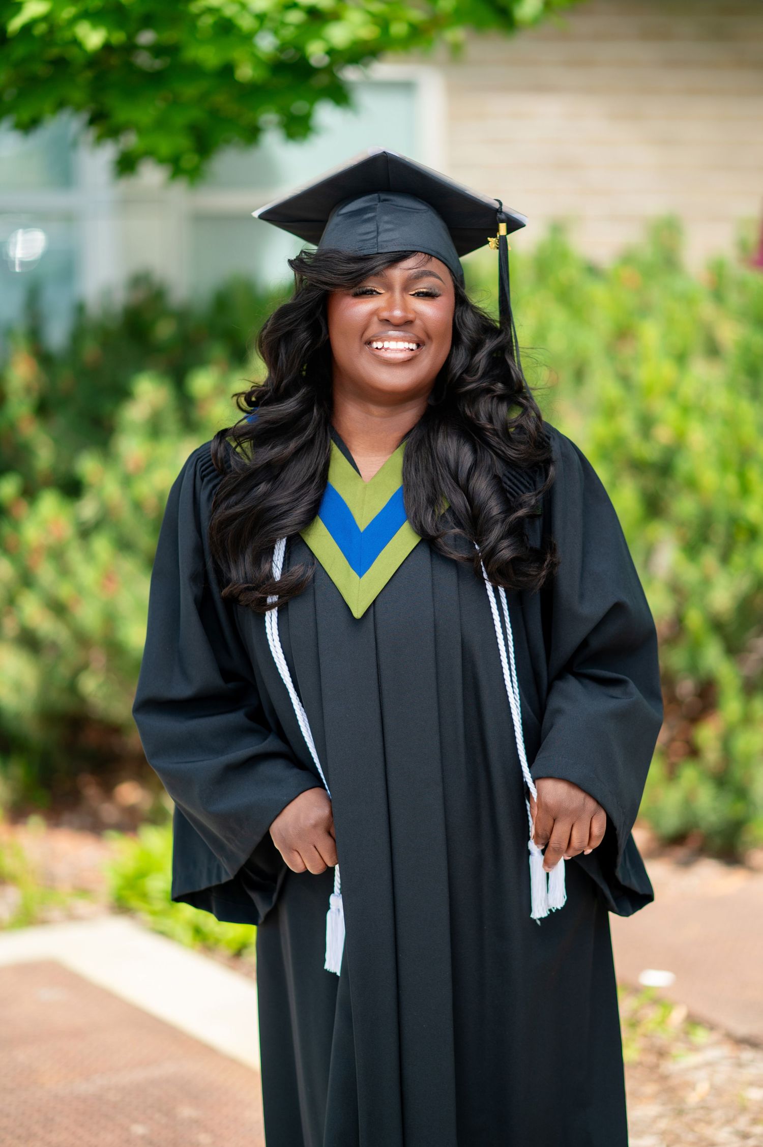 Graduate in traditional academic regalia poses with enthusiasm against garden background.