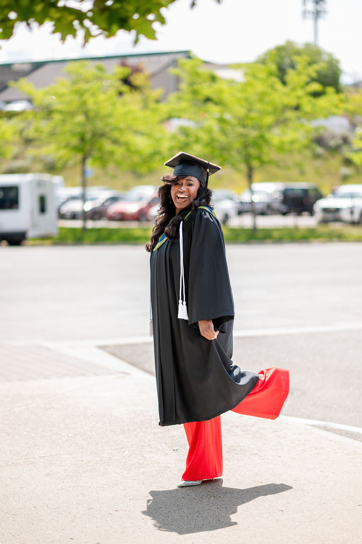 Graduate in red pants and black graduation gown poses playfully in outdoor setting.