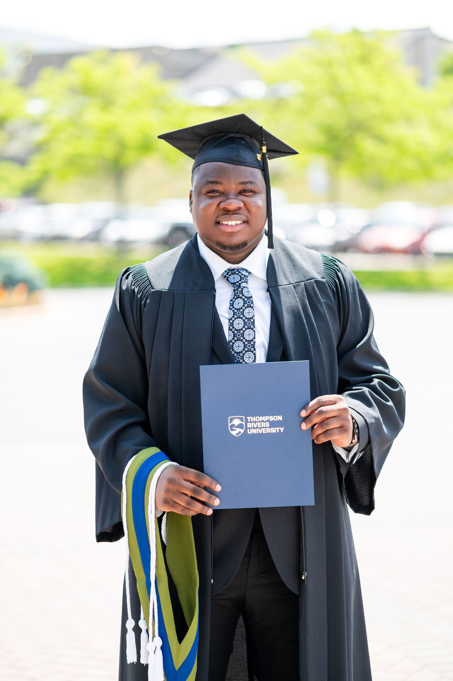 Graduate proudly displays diploma holder in black academic regalia during outdoor ceremony.