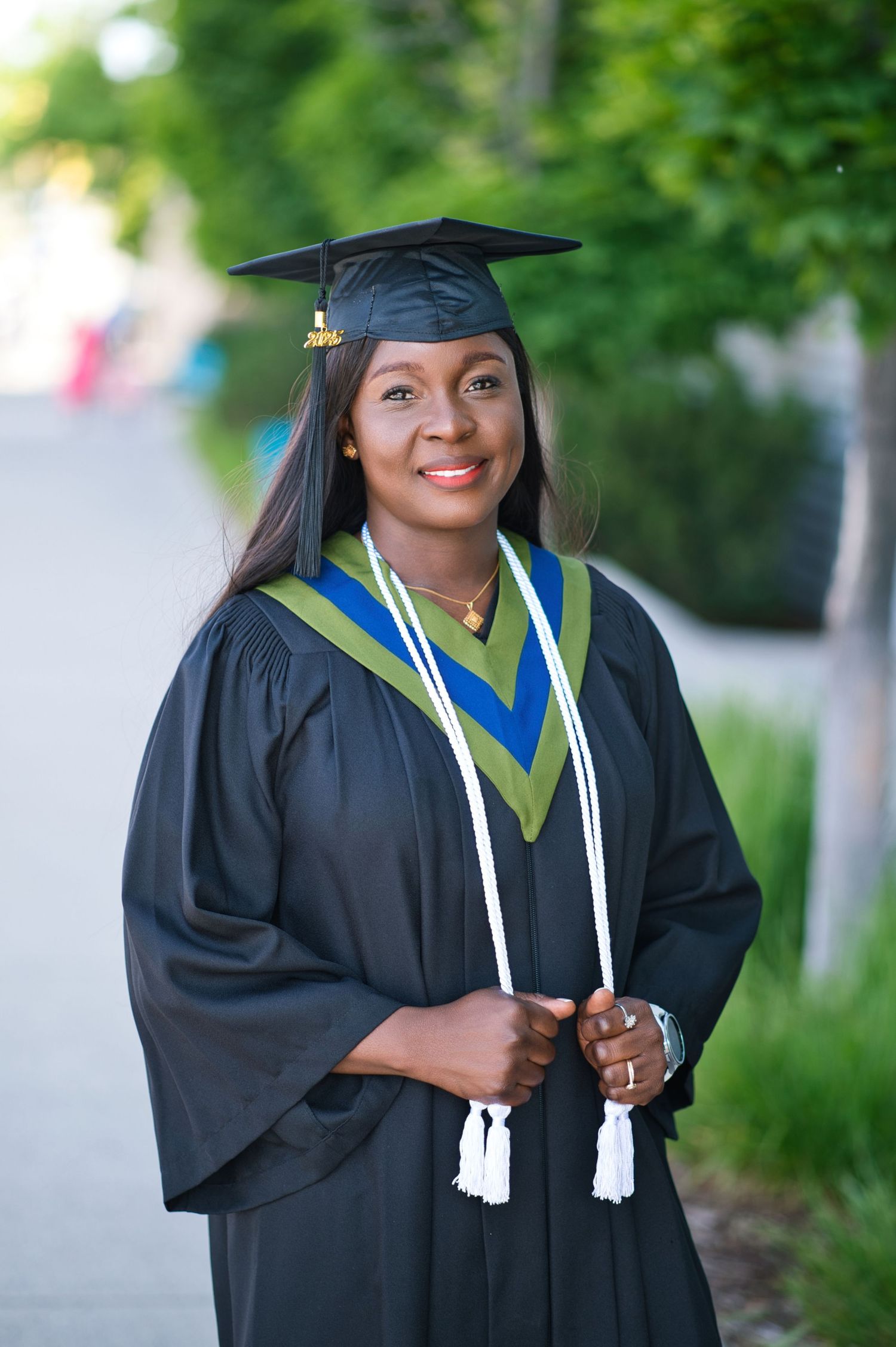 Graduate in academic dress stands confidently against lush green background.