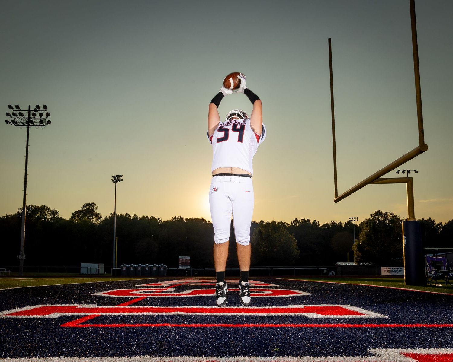 Football player stands alone on field at sunset with stadium lights and goal posts visible in background.