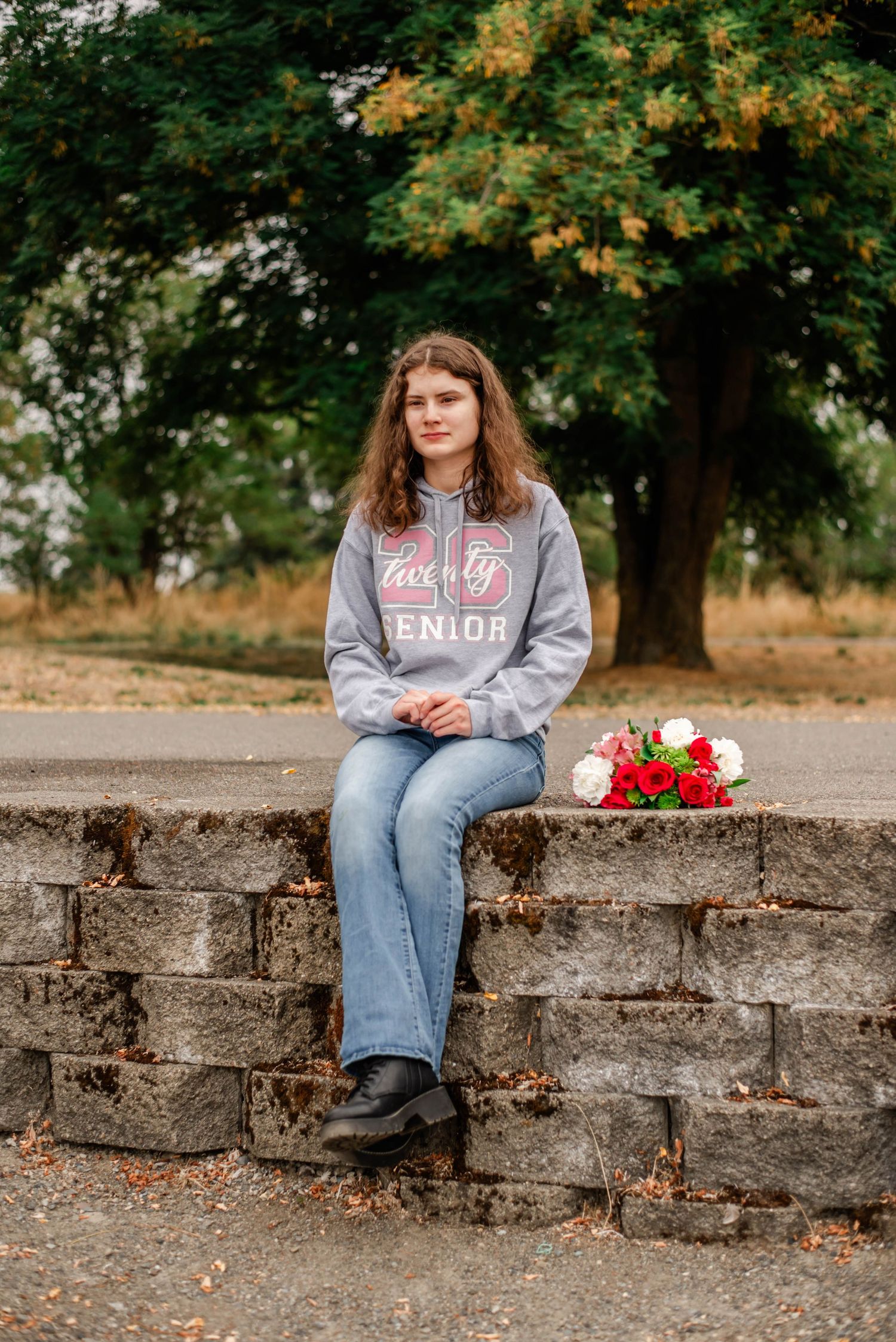 Person in gray sweatshirt and jeans sits on stone wall surrounded by autumn trees with flowers and clock as props.
