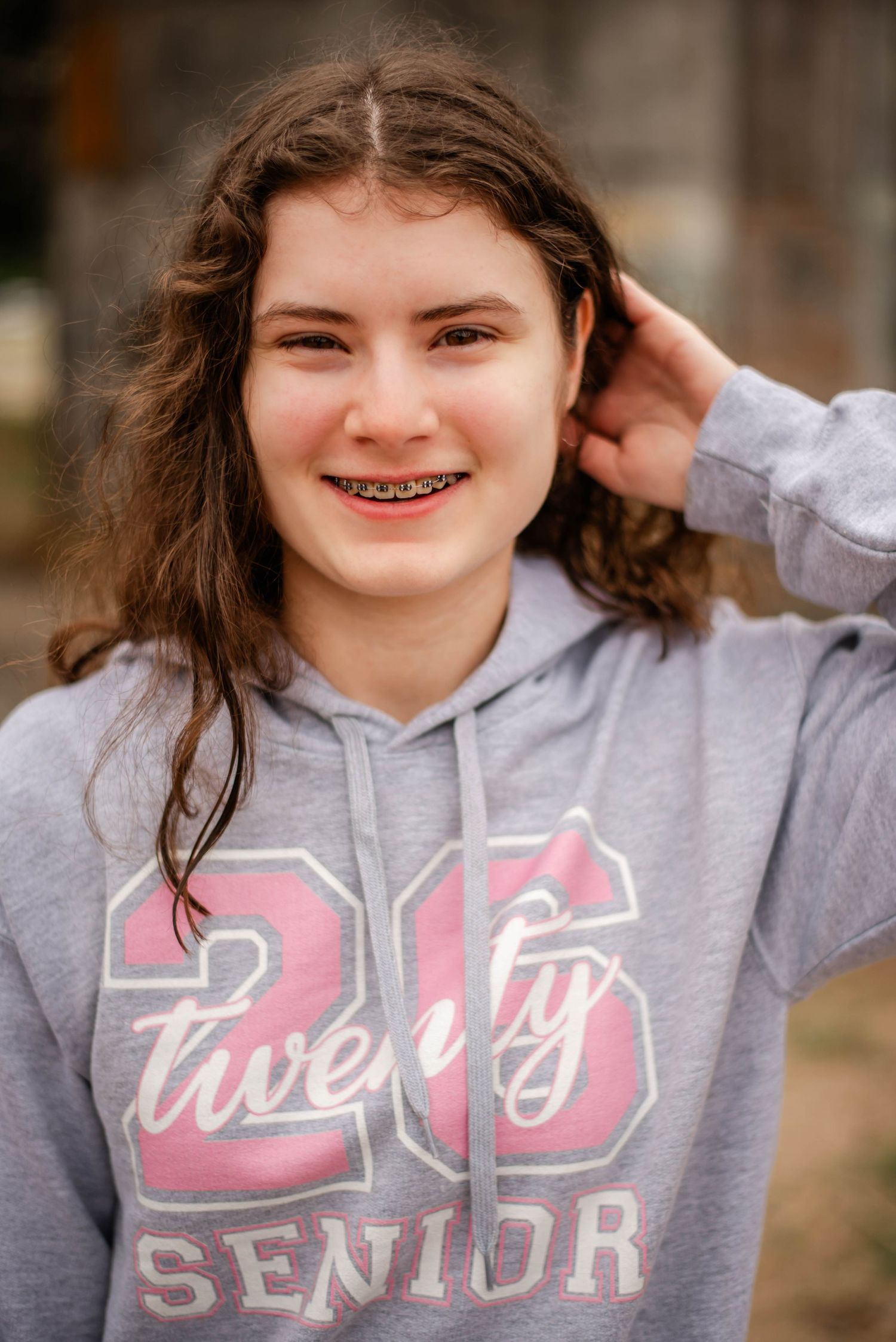 A student wearing a gray '2022 Senior' hooded sweatshirt smiles while touching their wavy brown hair outdoors.