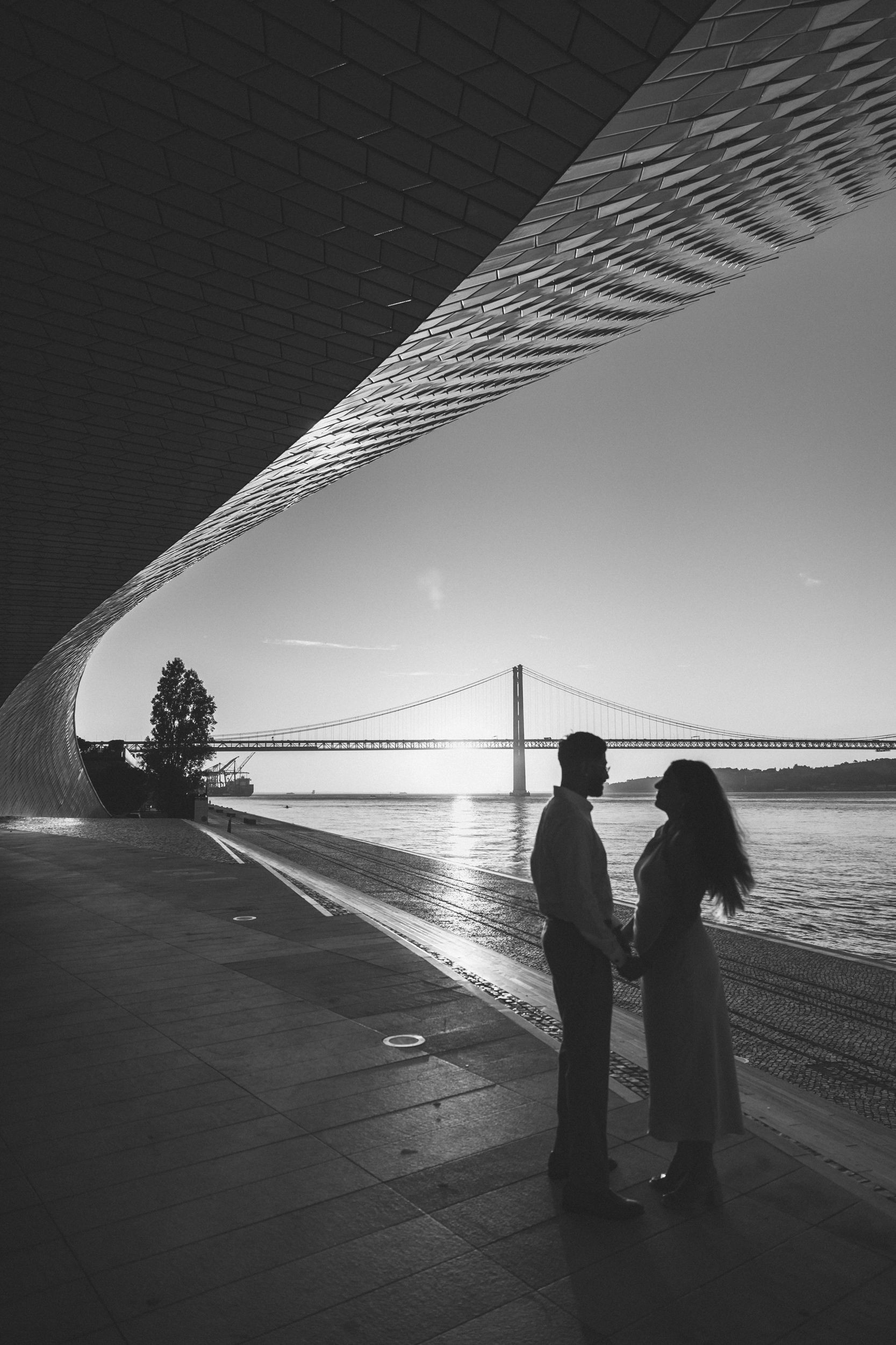 Candid moment of couple during sunrise engagement session with view of Lisbon’s 25 de Abril Bridge. Taken by a Portugal Wedding Photographer