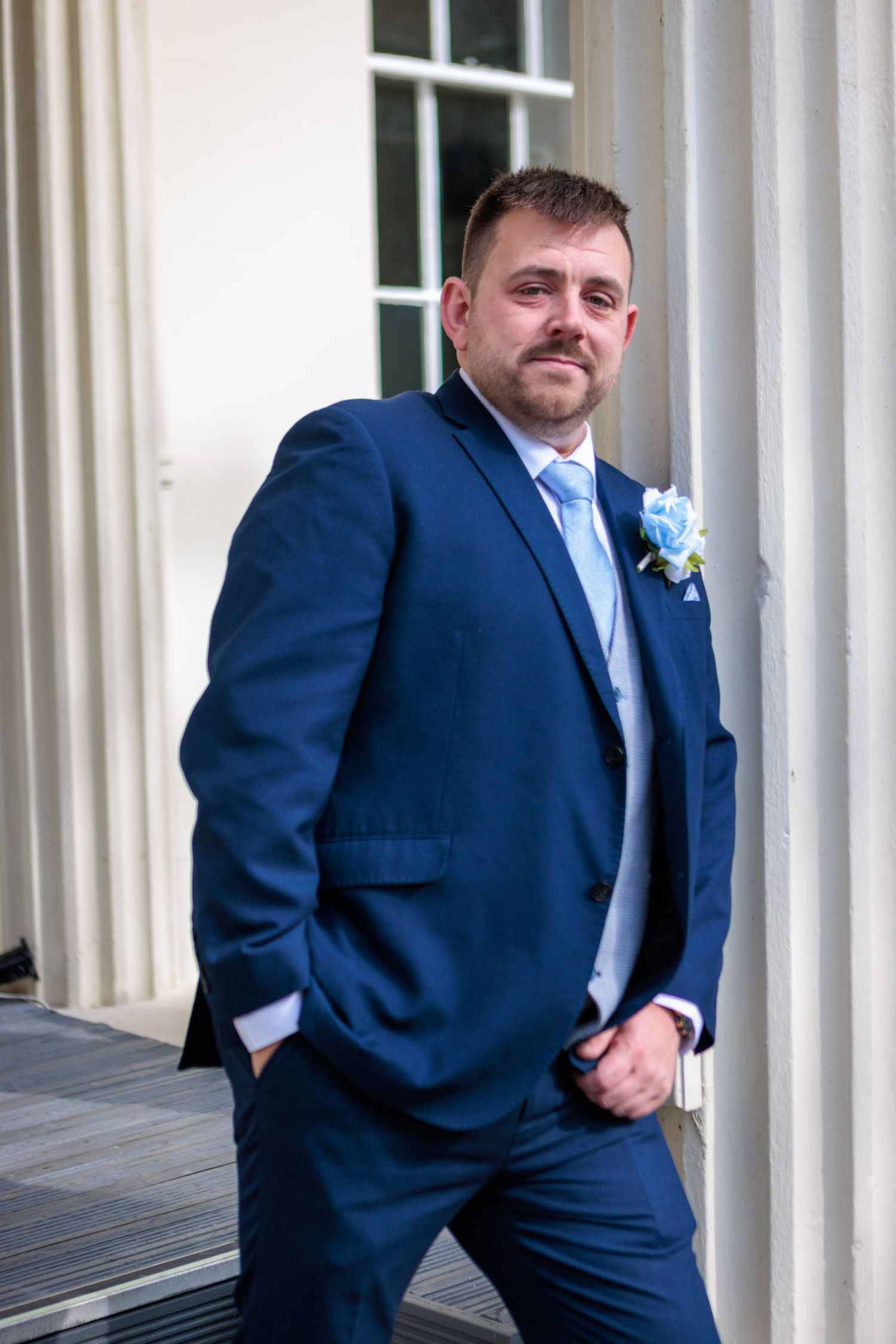 Man in navy blue suit poses confidently against white pillars in a formal setting.
