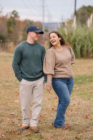 A couple wearing casual sweaters and jeans holds hands while walking together through a grassy autumn field.