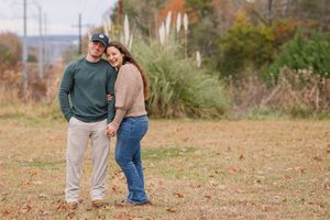 A couple in casual fall attire embraces while standing in a grassy field with autumn foliage in the background.
