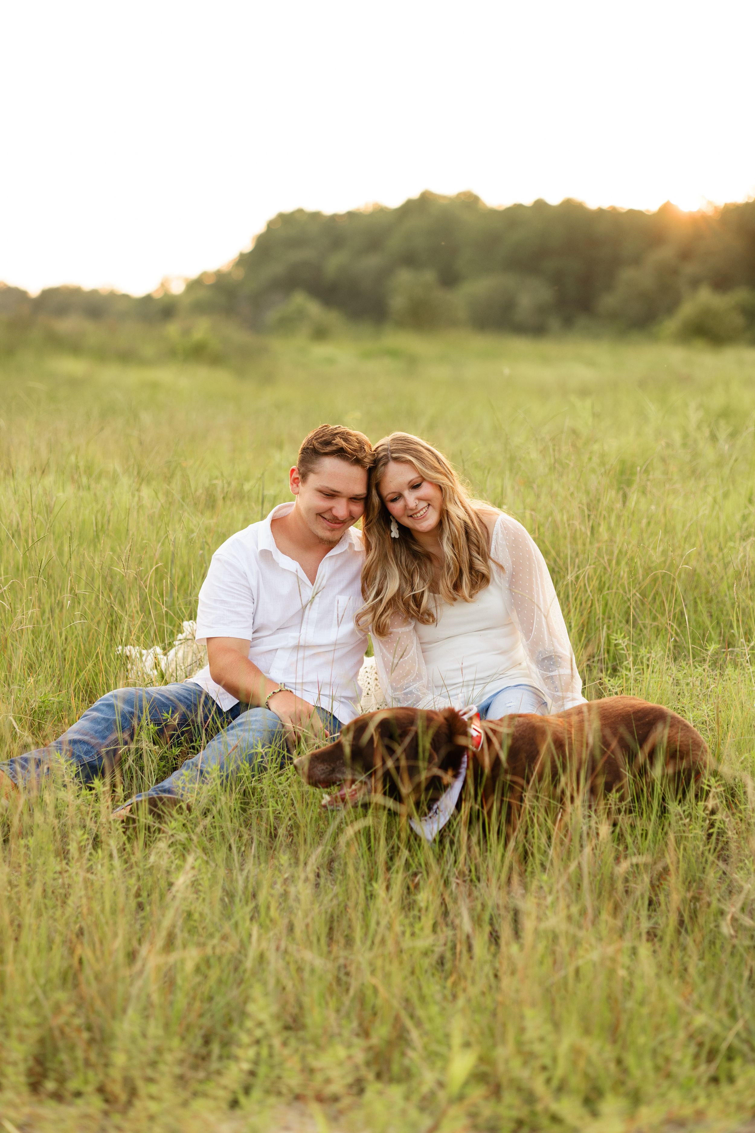 A couple and their dog relax together in a sunlit grassy meadow during golden hour.