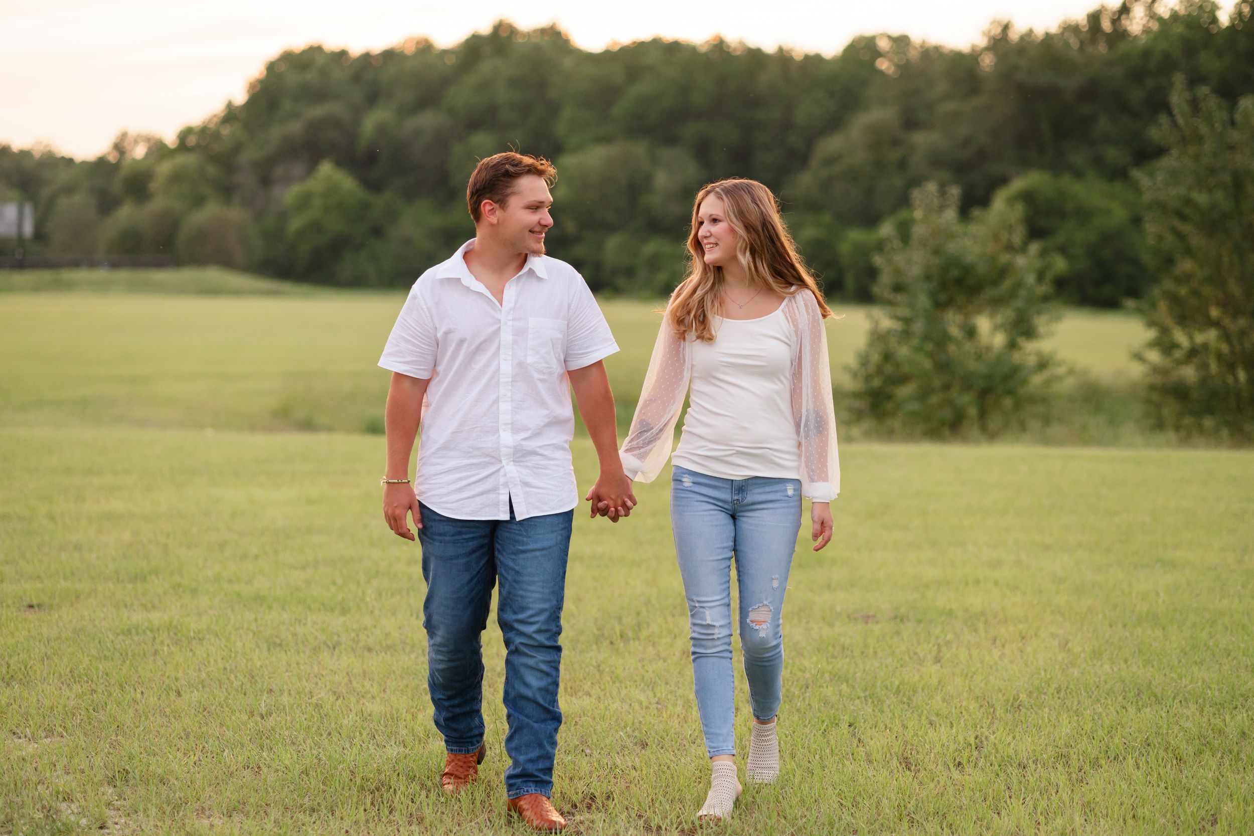 Couple walking hand in hand through a serene meadow with trees in the background.