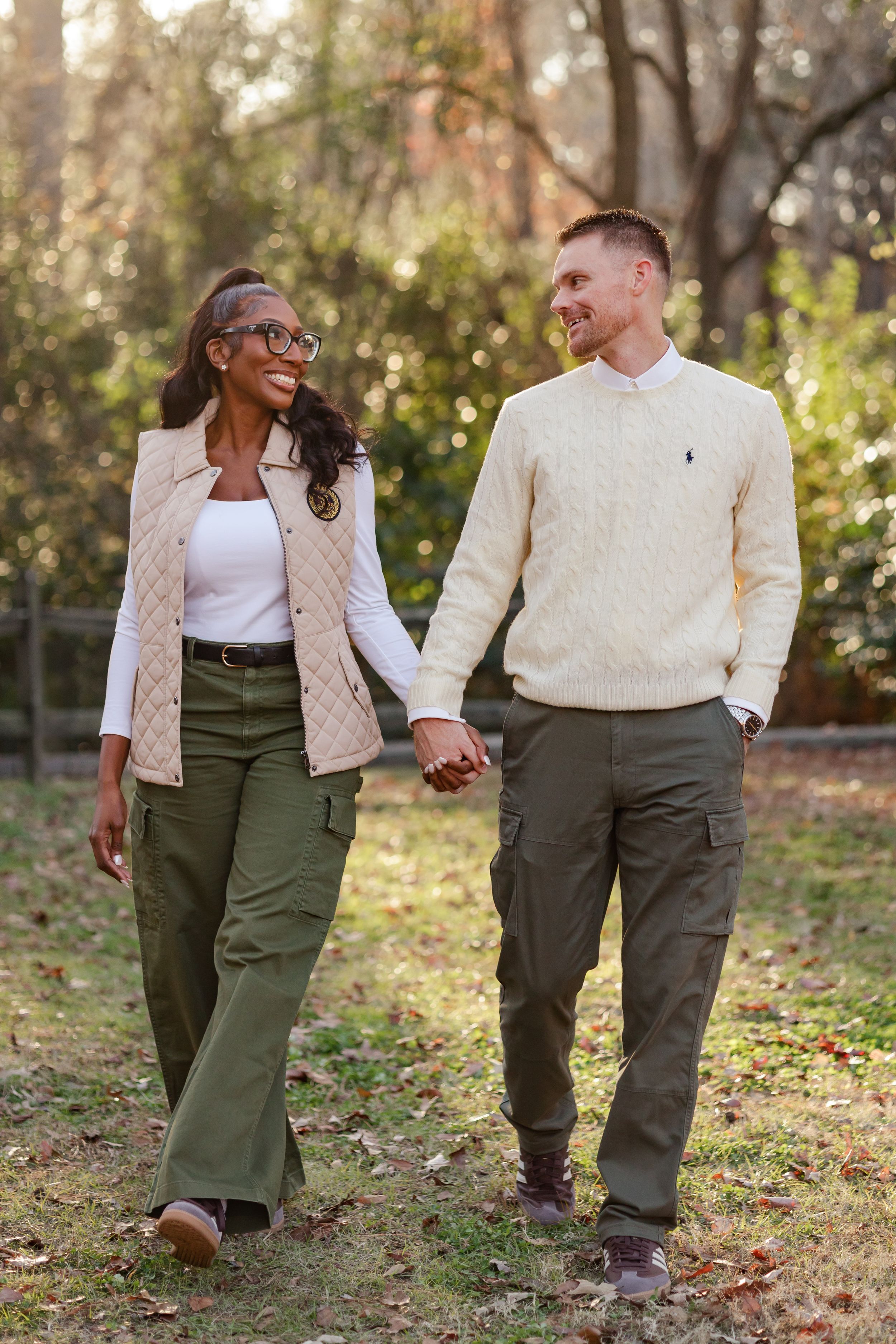 A couple in coordinated outfits with beige jackets and olive pants walk together through fallen autumn leaves in a park.