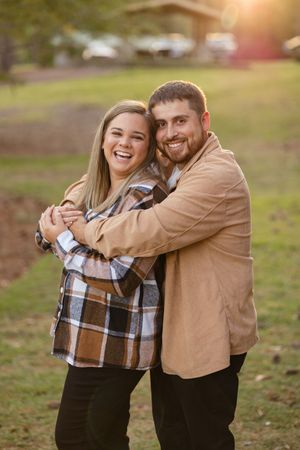 A happy couple embraces outdoors during golden hour, wearing casual fall attire including plaid and corduroy.