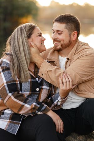 A couple shares tender moments by a lake at sunset while wearing casual clothing and a plaid flannel.