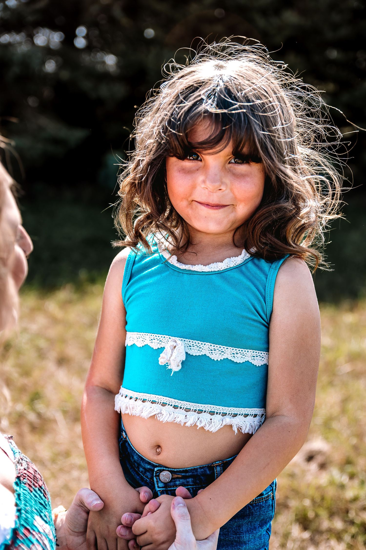 A young child in a turquoise crop top and denim shorts plays outdoors in a sunlit meadow.
