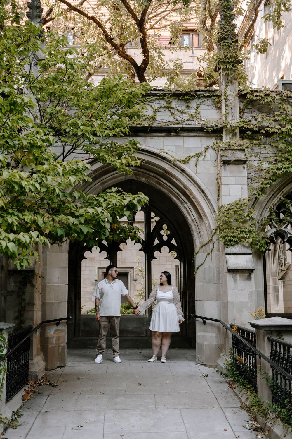 A couple walks through a Gothic stone archway covered in green ivy at a university campus.