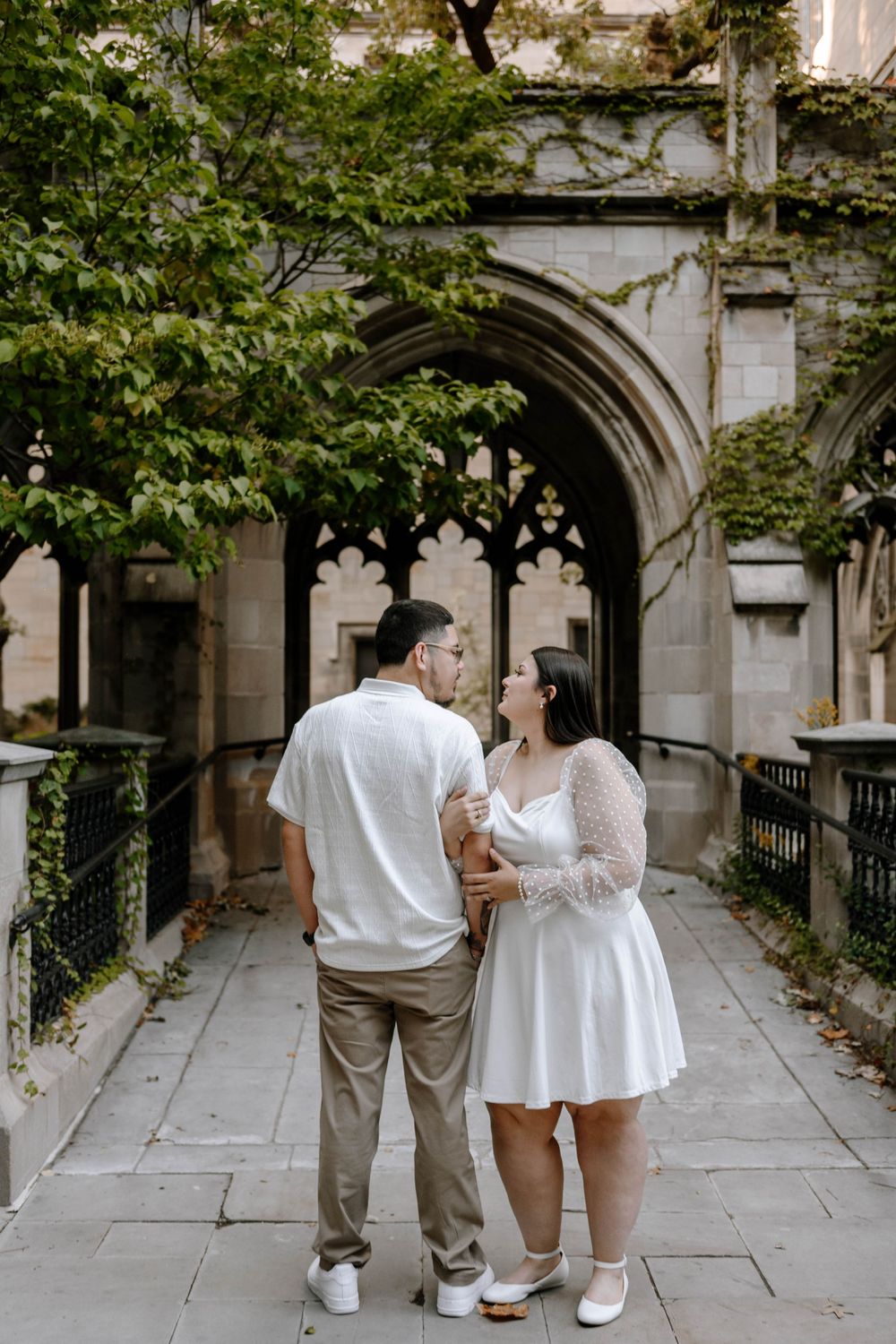 A couple embraces in a beautiful stone passageway with gothic architectural details and green foliage.