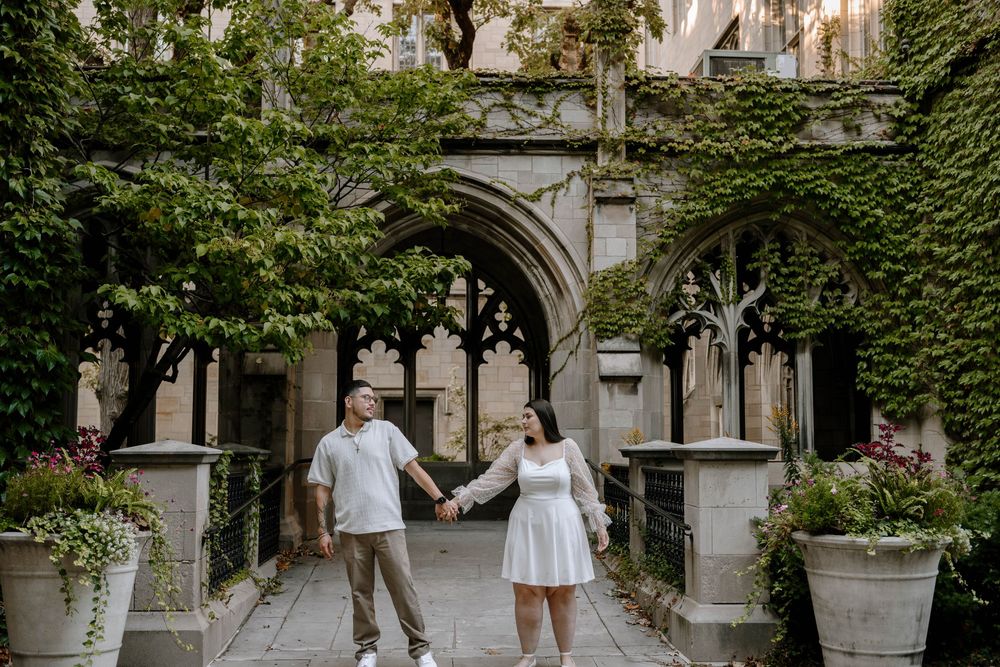A couple holds hands while walking through an ivy-covered Gothic archway in a romantic courtyard setting.
